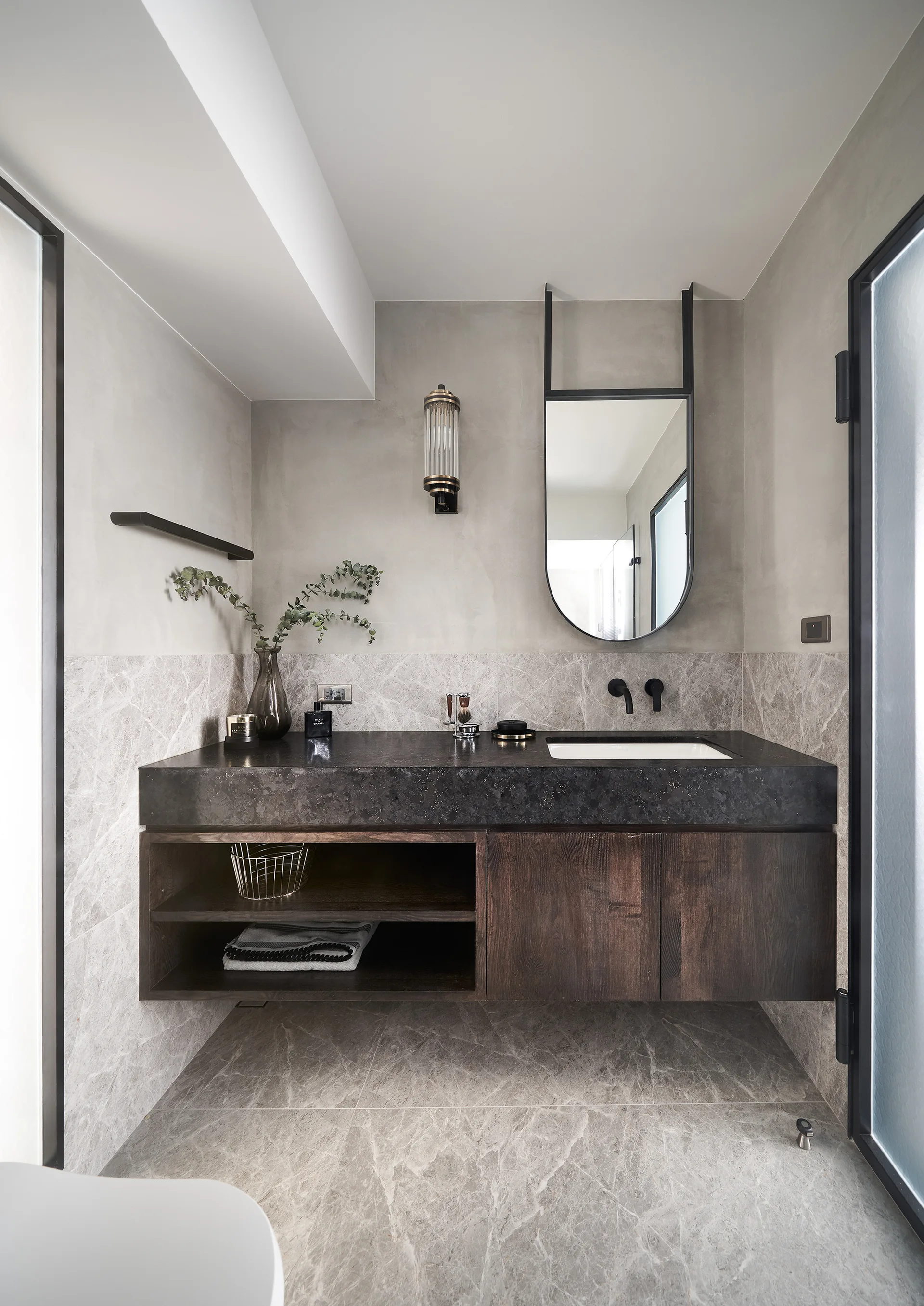 A modern grey bathroom featuring a floating dark wood vanity with a black stone countertop, an oval mirror, and veined stone floor tiles.