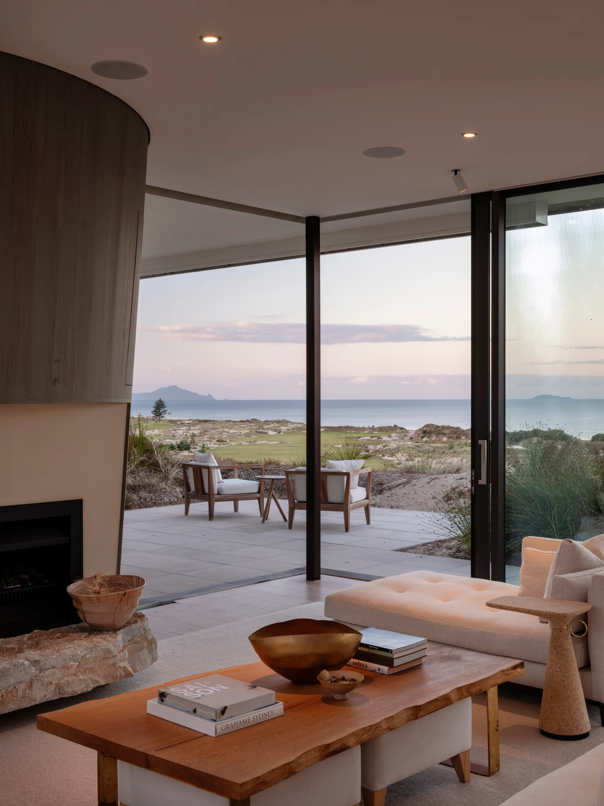 An organic modern living room featuring a stone hearth fireplace, wooden coffee table, and large glass doors opening to a patio with ocean views.