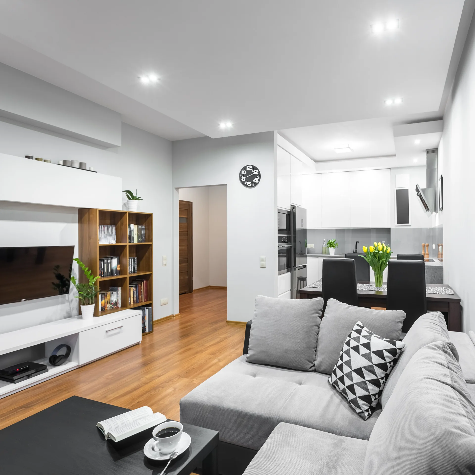 A contemporary open plan living room features a grey sectional sofa, a white media console with a wood bookshelf, and an adjoining white kitchen.