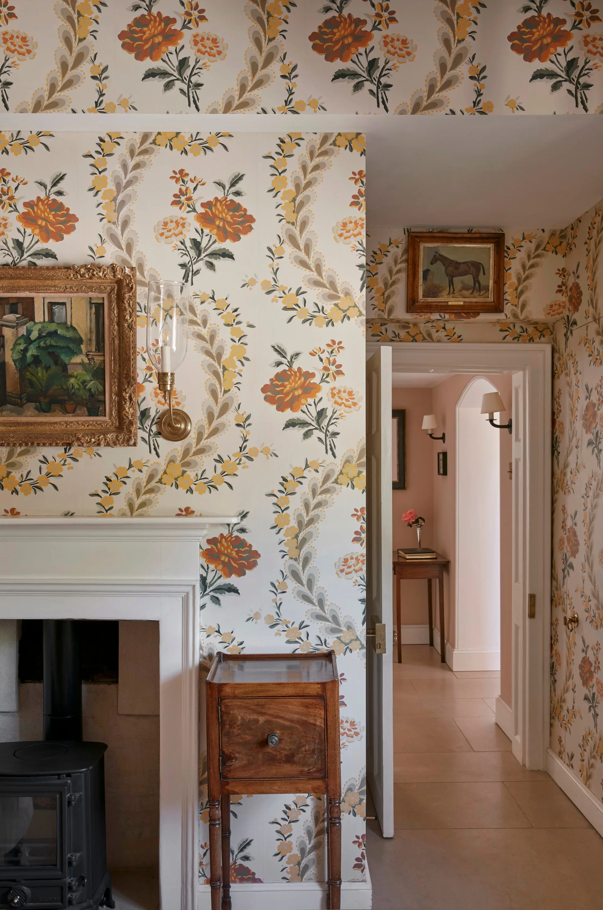 A traditional living room features orange floral wallpaper, a white fireplace mantel with a wood stove, and a view into a hallway.