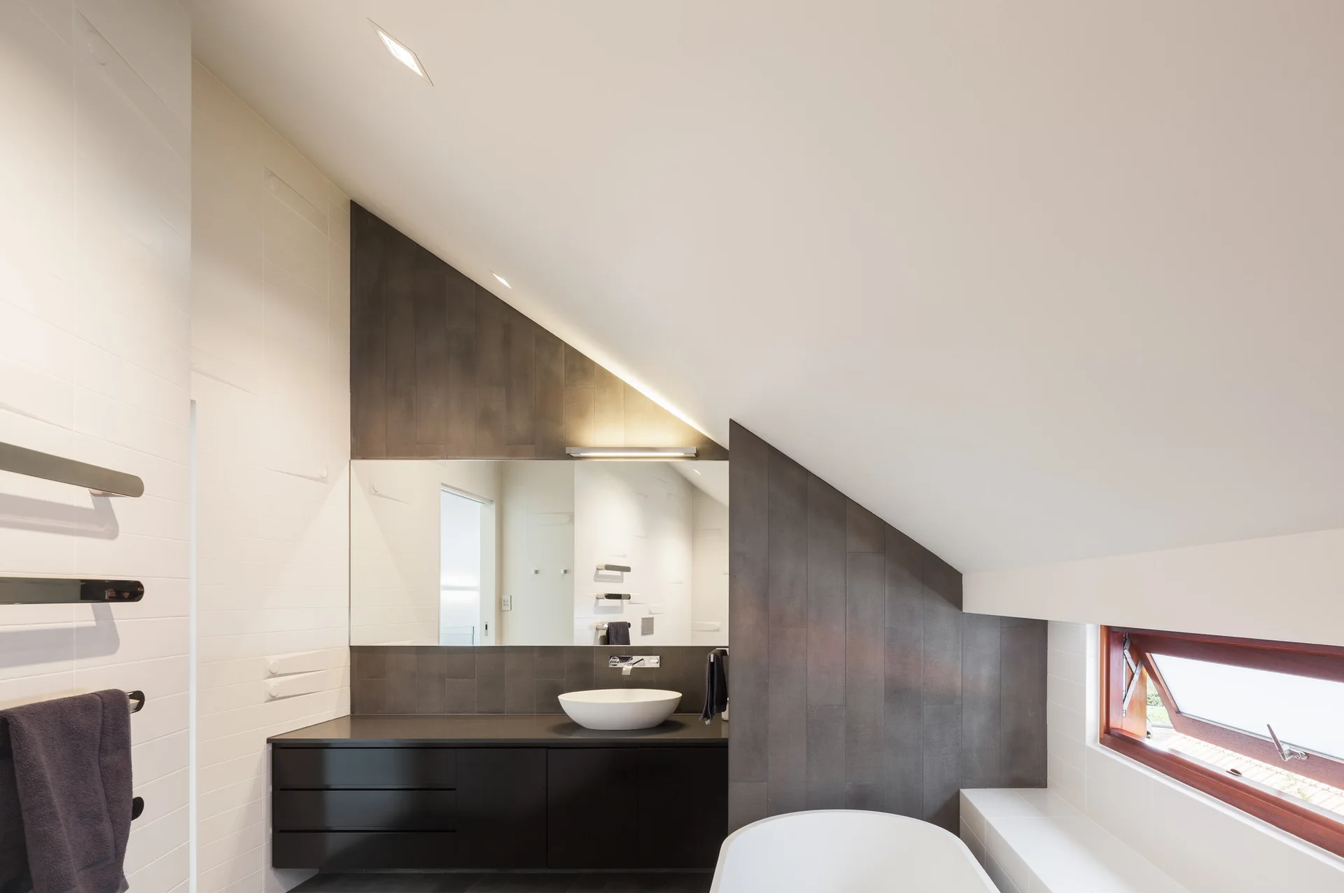 A modern attic bathroom features a black vanity with a white vessel sink, a large rectangular mirror, and dark grey tile on a sloped wall.