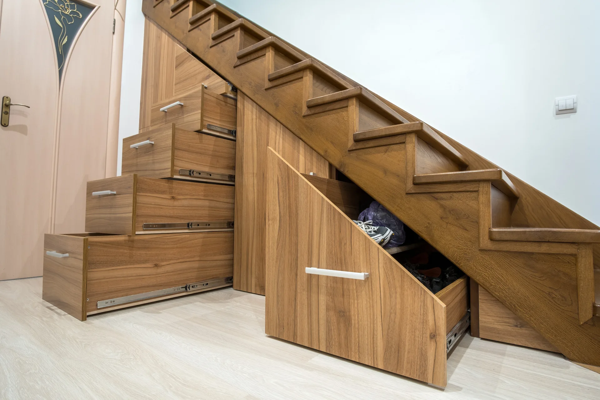 A wooden staircase features several open pull-out storage drawers integrated into the under-stair space on light-colored laminate flooring.