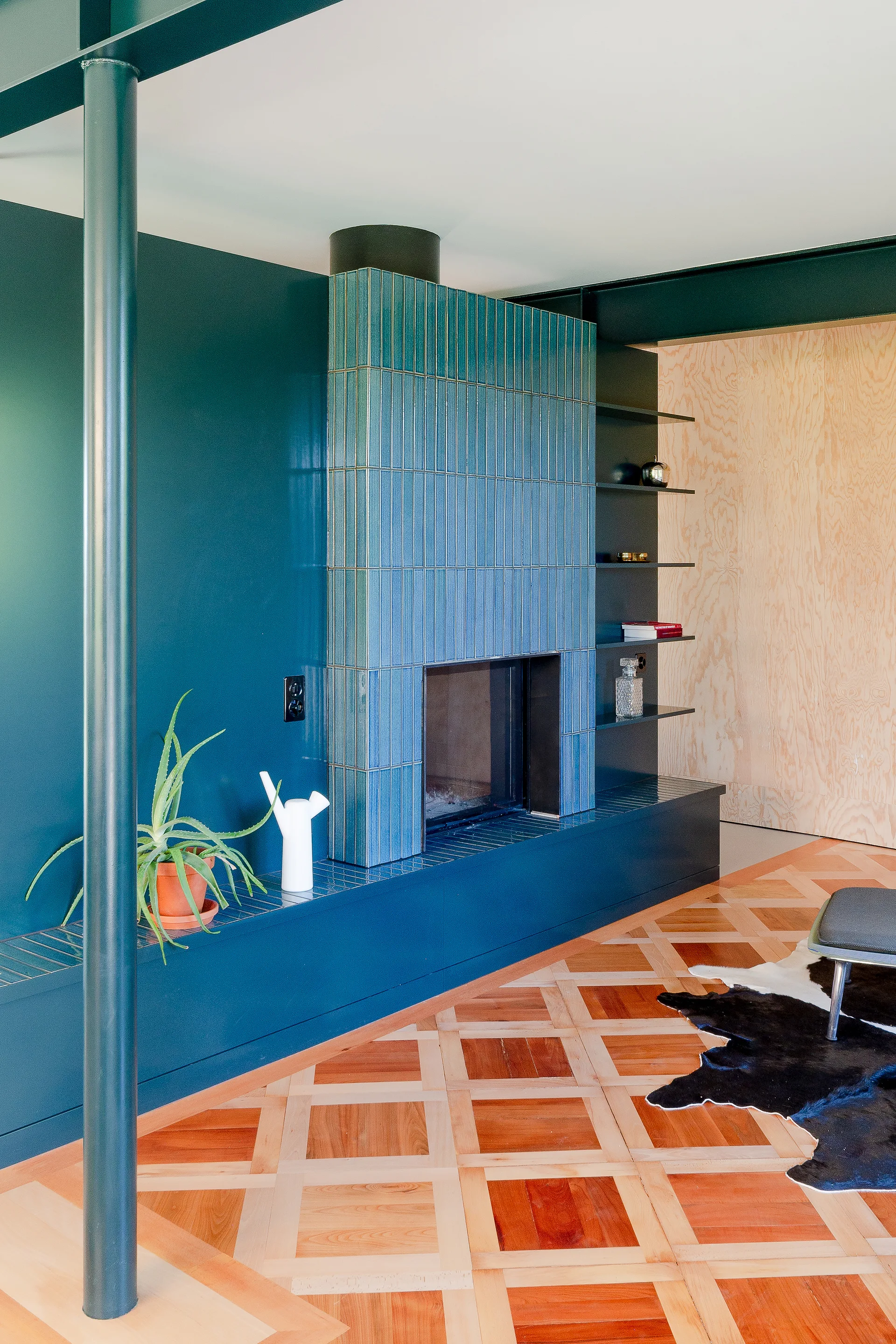 A modern living room features a blue vertical-tile fireplace, deep teal walls, a dark metal column, and decorative geometric wood parquet flooring.