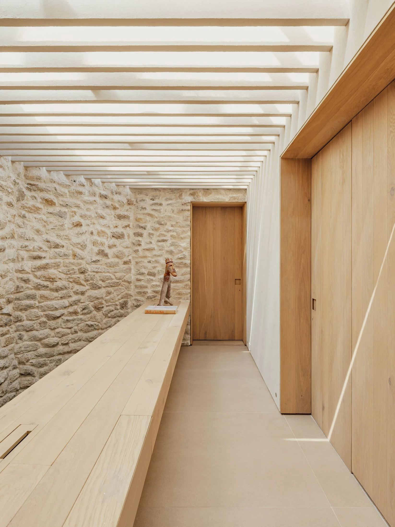 A minimalist hallway featuring a textured stone wall on one side, light wood wall panels on the other, and a slatted ceiling casting striped shadows.