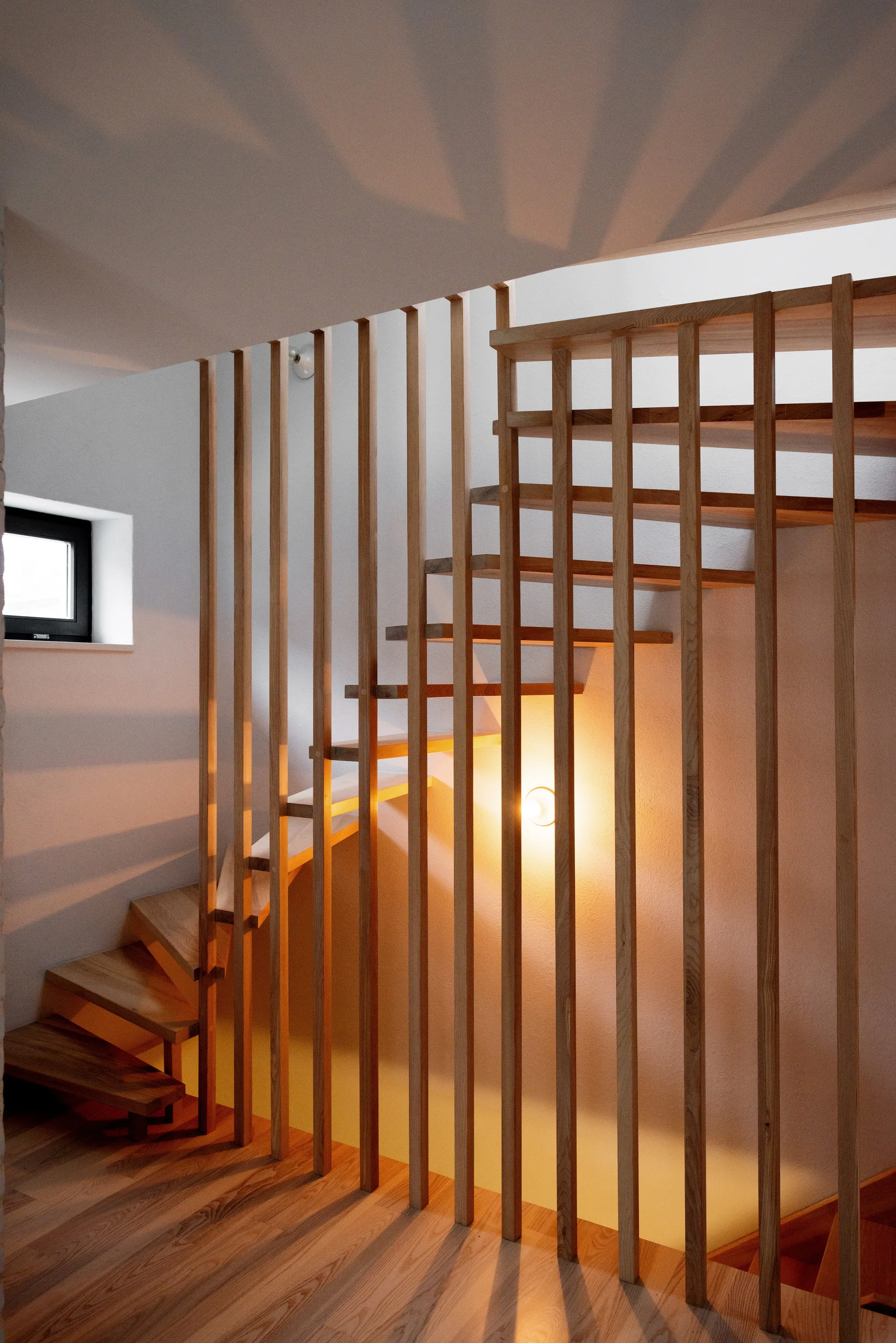 A minimalist wooden staircase features vertical wood slats serving as a balustrade, illuminated by a warm round wall sconce.