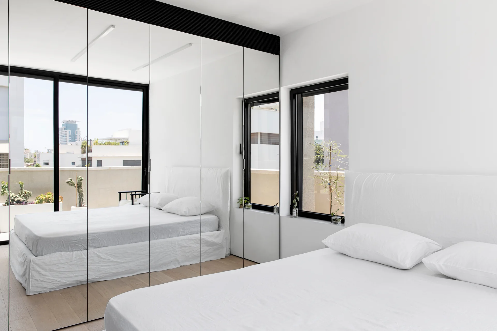 A minimalist bedroom features a white upholstered bed with matching linens next to a wall of floor-to-ceiling mirrored closet doors.