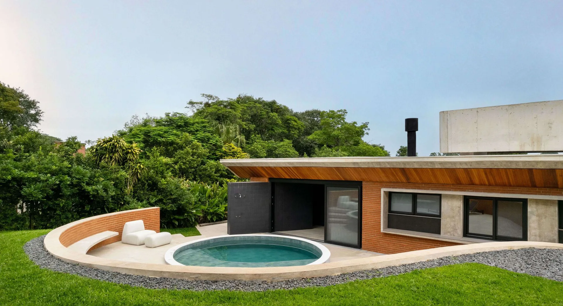 A circular pool is integrated into a paved patio alongside a modern brick and concrete house with a curved brick retaining wall.