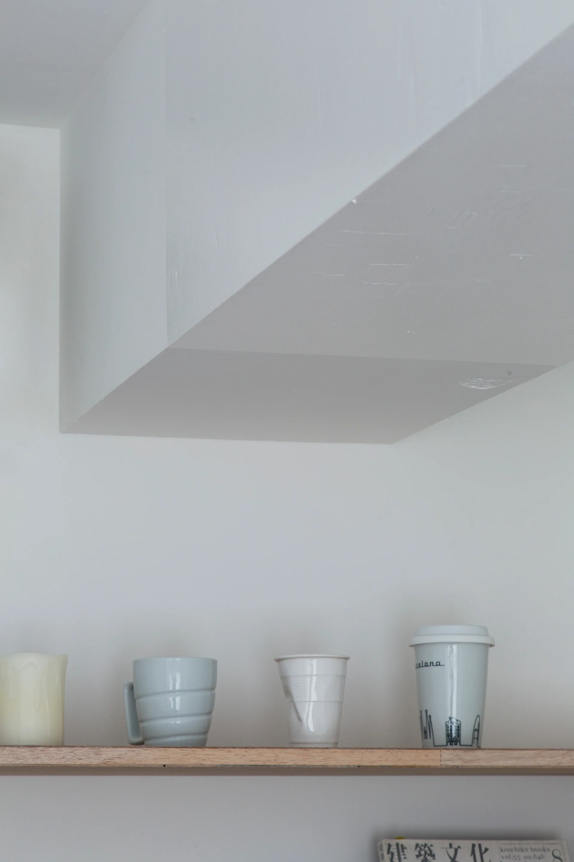 A close-up of a natural wood shelf mounted on a white wall underneath a painted soffit, holding ceramic mugs and a wax candle.