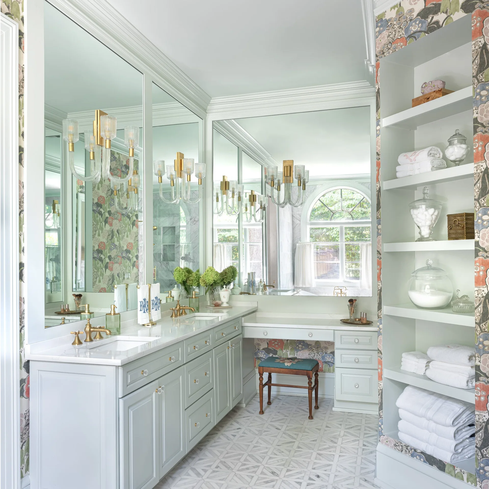 A traditional bathroom featuring a pale blue double vanity, large mirrors with brass sconces, and open shelving styled with towels.