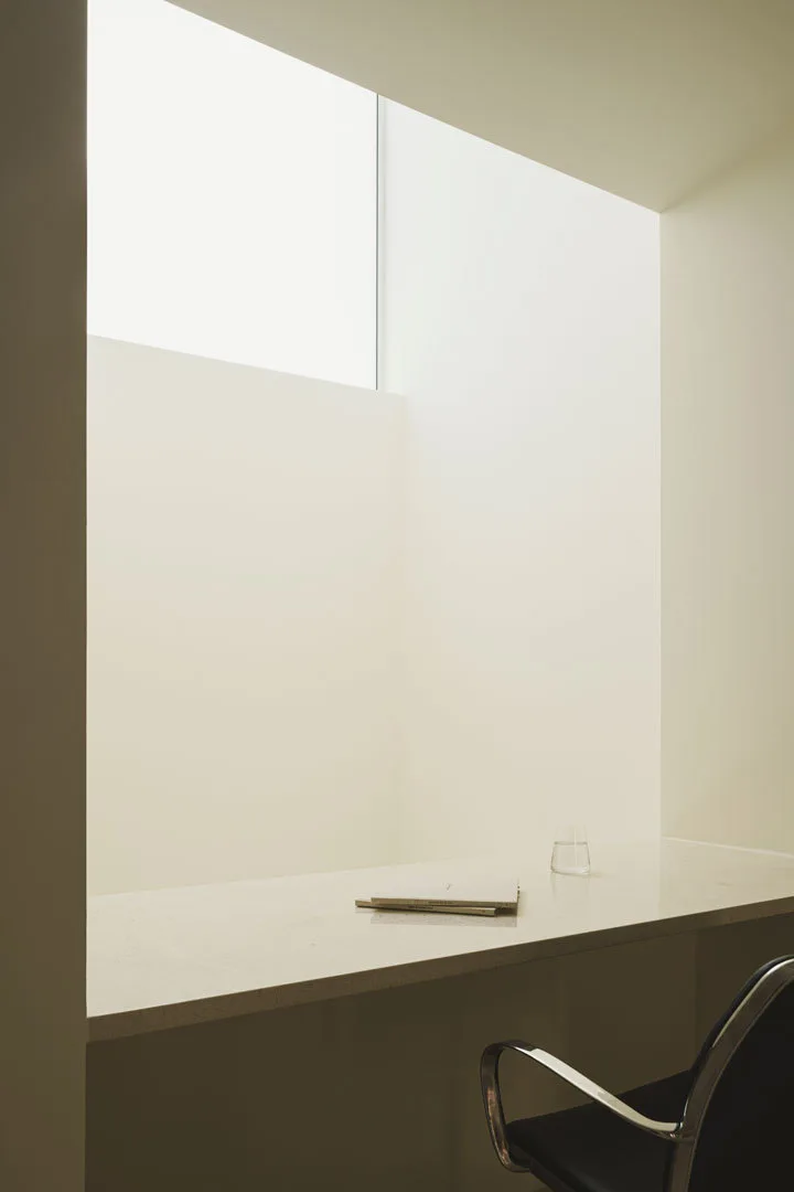 A minimalist off-white study nook features a stone-like desk built beneath a large ceiling skylight next to a black and chrome chair.
