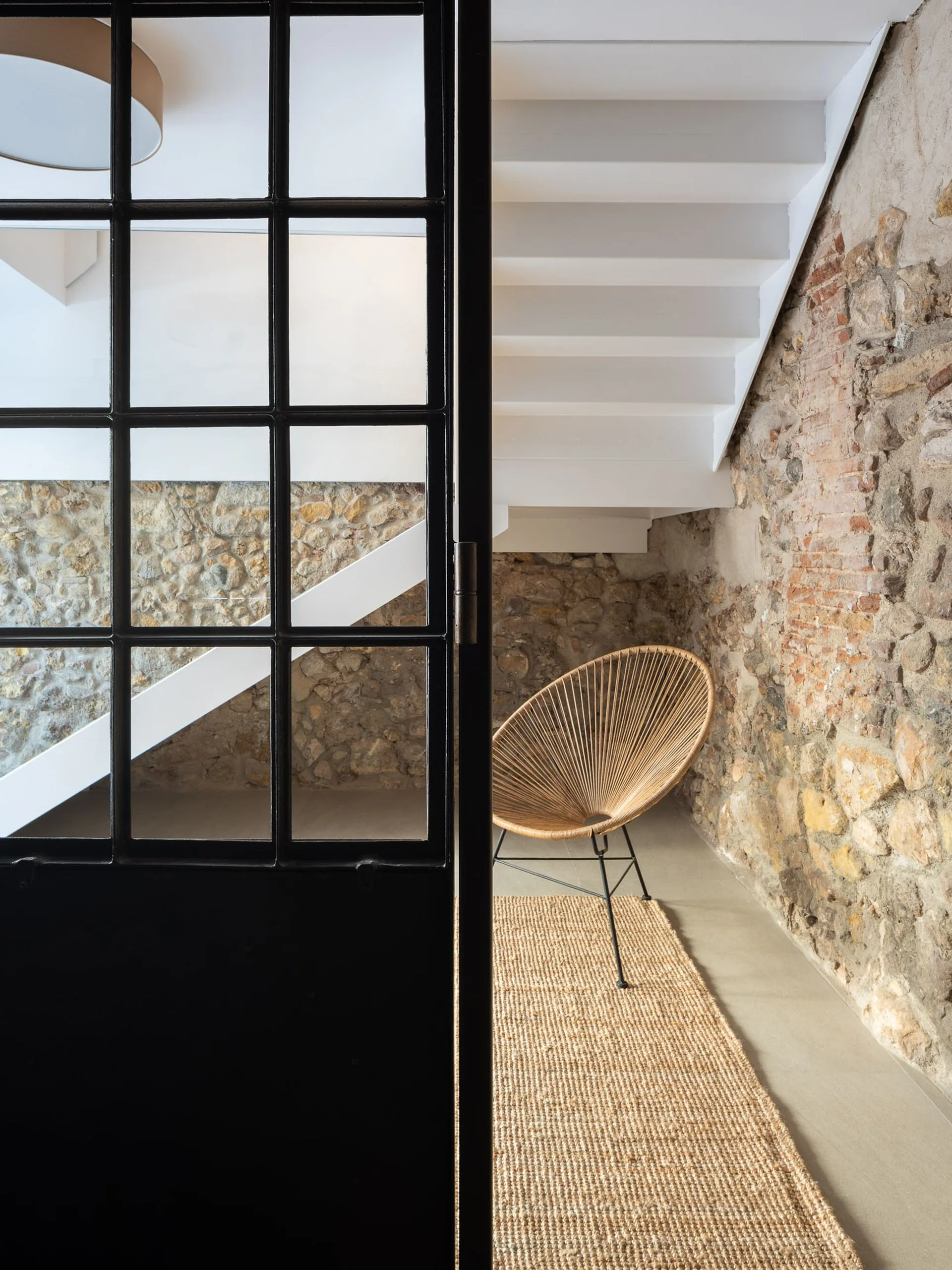 A view through a black metal framed glass door showing white stairs, an exposed stone wall, a woven bowl-style chair, and a jute rug.