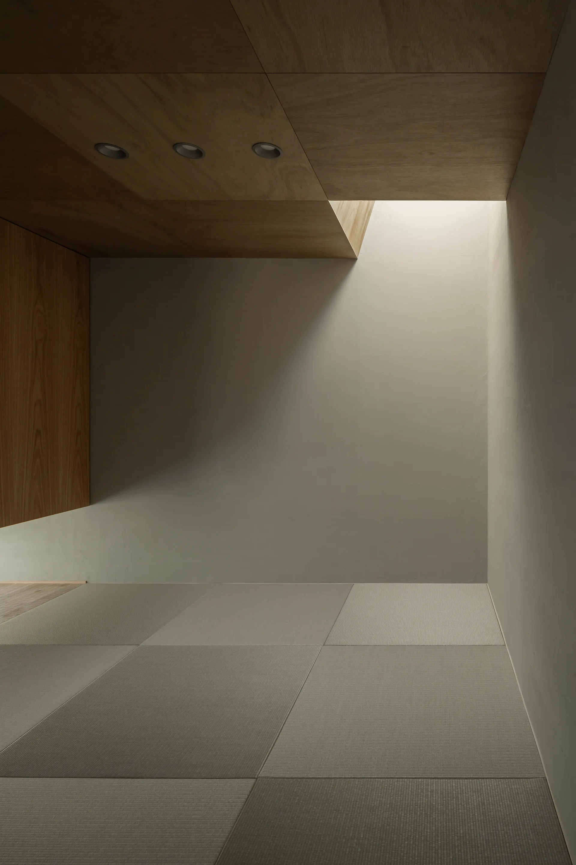 A minimalist Japanese room features gray tatami floor mats, smooth plaster walls, and a warm wood-paneled ceiling with a skylight.