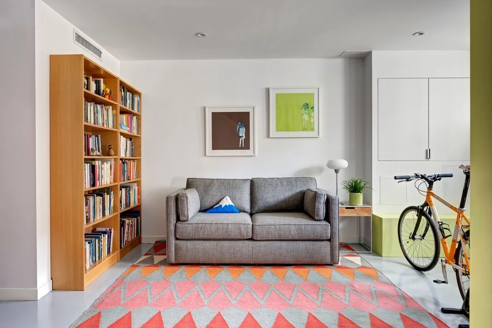A grey fabric sofa sits on a colorful geometric rug next to a tall wooden bookshelf filled with books, with a bicycle parked on the right.