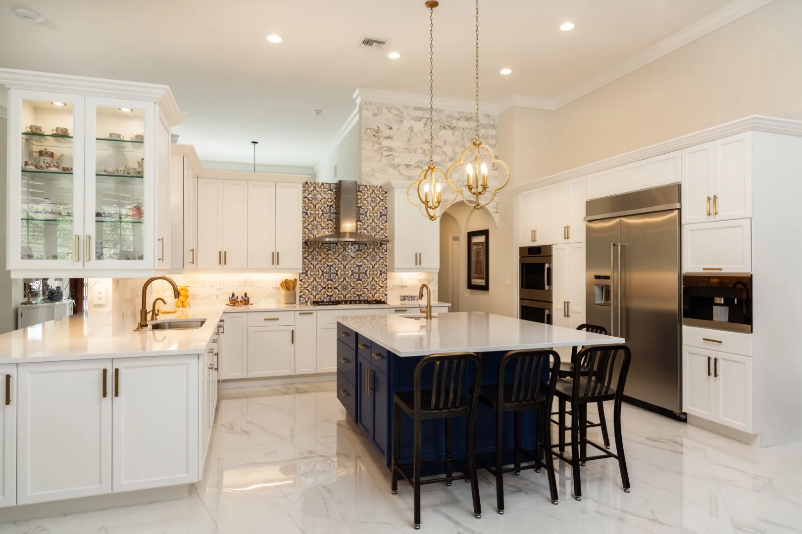 A bright kitchen features white perimeter cabinets, a blue central island with black barstools, gold pendant lights, and polished marble flooring.