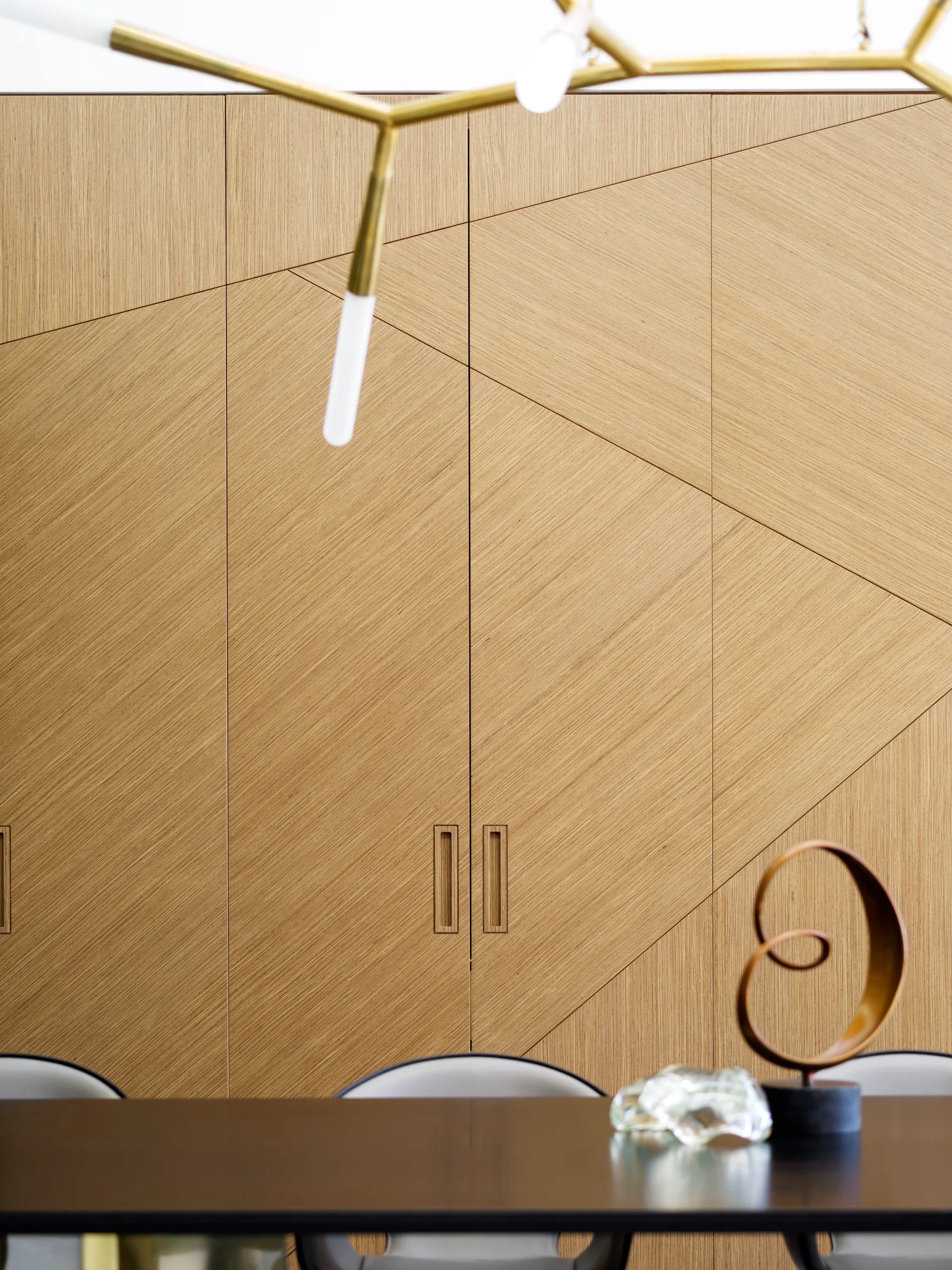 A wall of custom wood cabinetry with geometric diagonal seams stands behind a dark dining table decorated with a gold metal spiral sculpture.