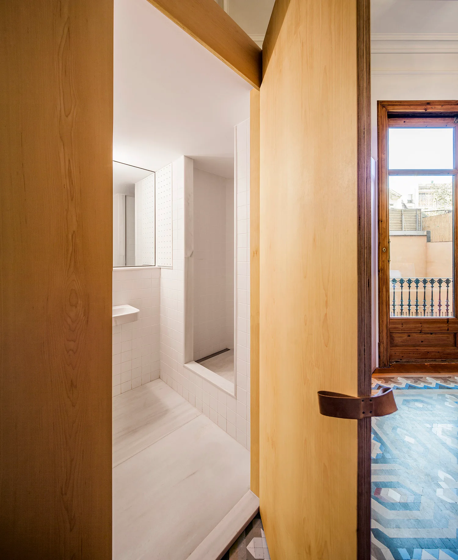 A view through an open light wood plywood door into a minimalist white-tiled bathroom with a geometric patterned tile floor in the foreground.