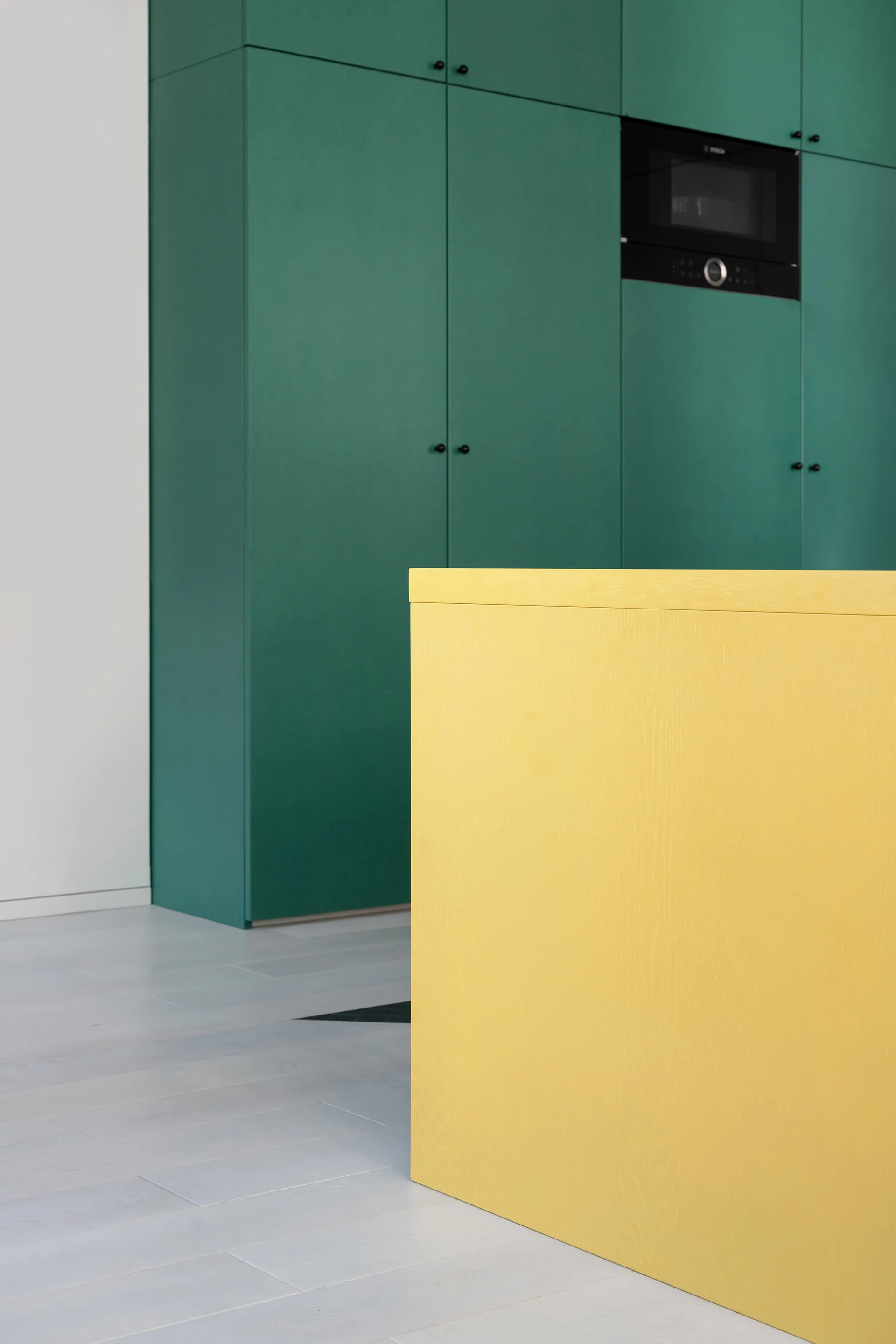 A contemporary kitchen featuring flat-panel green cabinetry with a built-in oven alongside a bright yellow wooden island and light wood plank flooring.