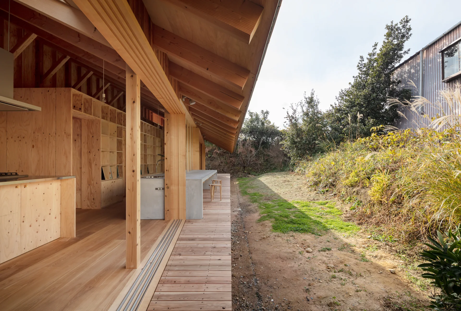 A minimalist open-plan wood kitchen with a concrete island opens directly onto a wood-planked deck next to a natural garden.