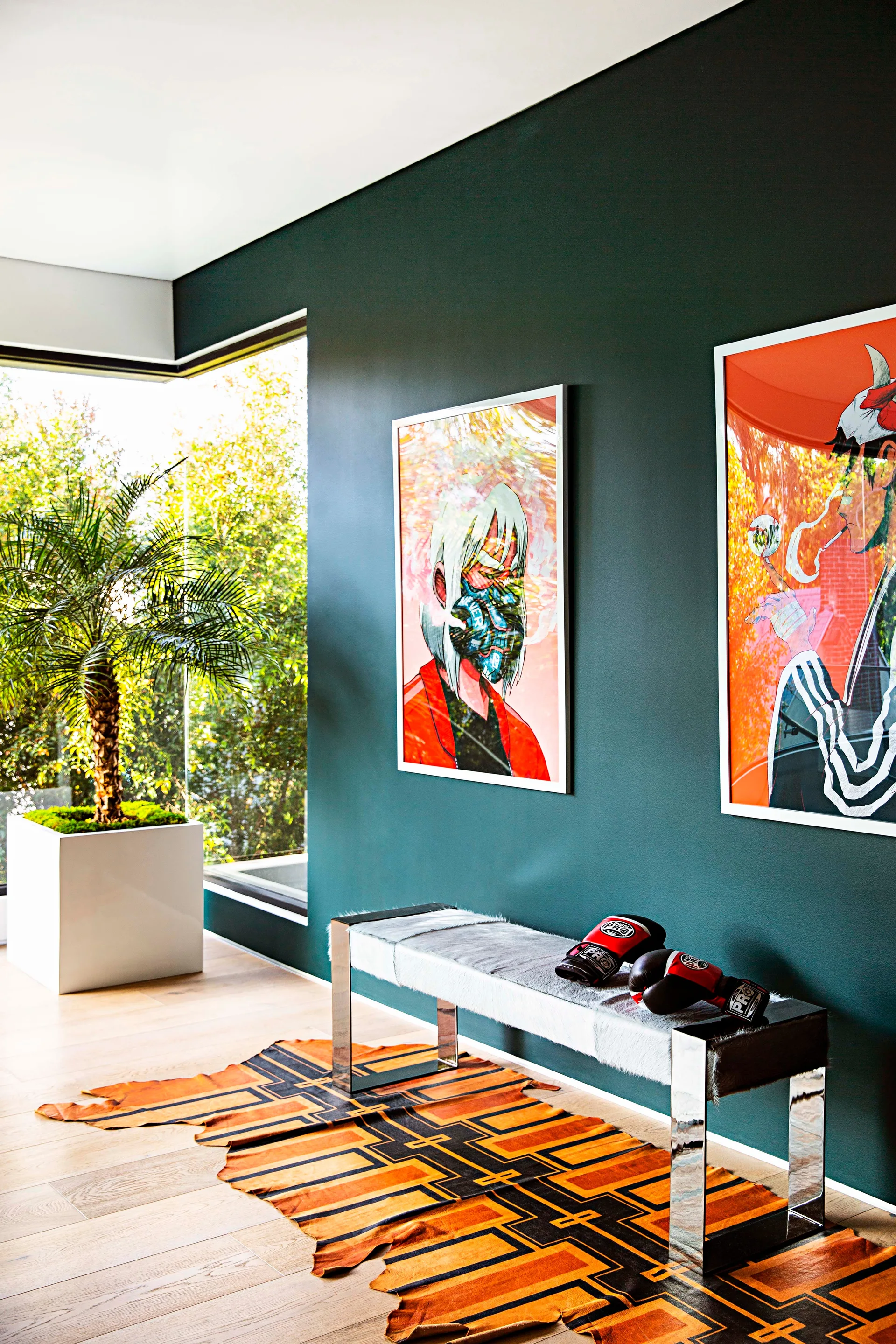 Two framed graphic art prints hang on a dark green wall above a chrome bench and a geometric orange hide rug on light wood floors.