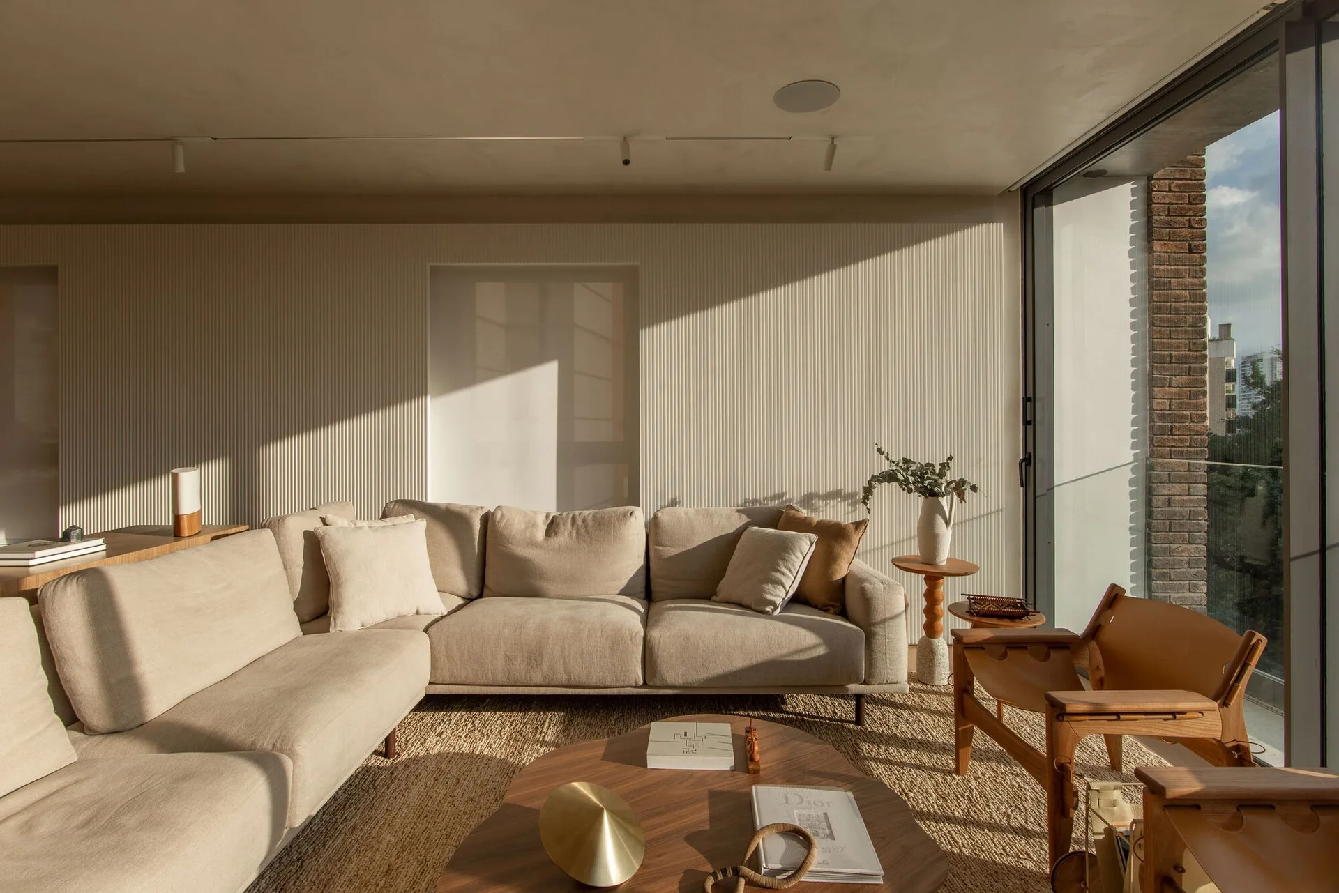 A minimalist living room features a beige sectional sofa, cream fluted wall panels, an organic wood coffee table, and a woven jute area rug.