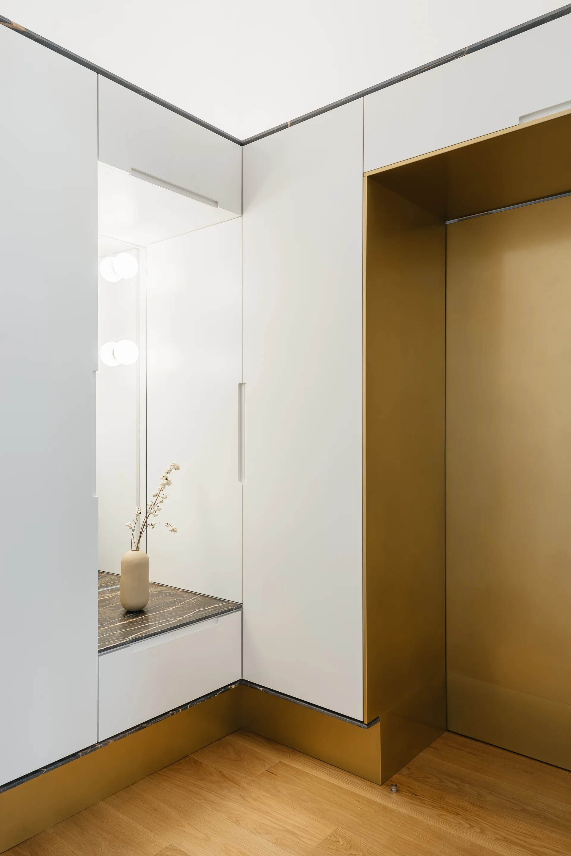 A built-in white wardrobe system features a recessed mirror vanity with a dark marble countertop, integrated round sconces, and a metallic gold base.