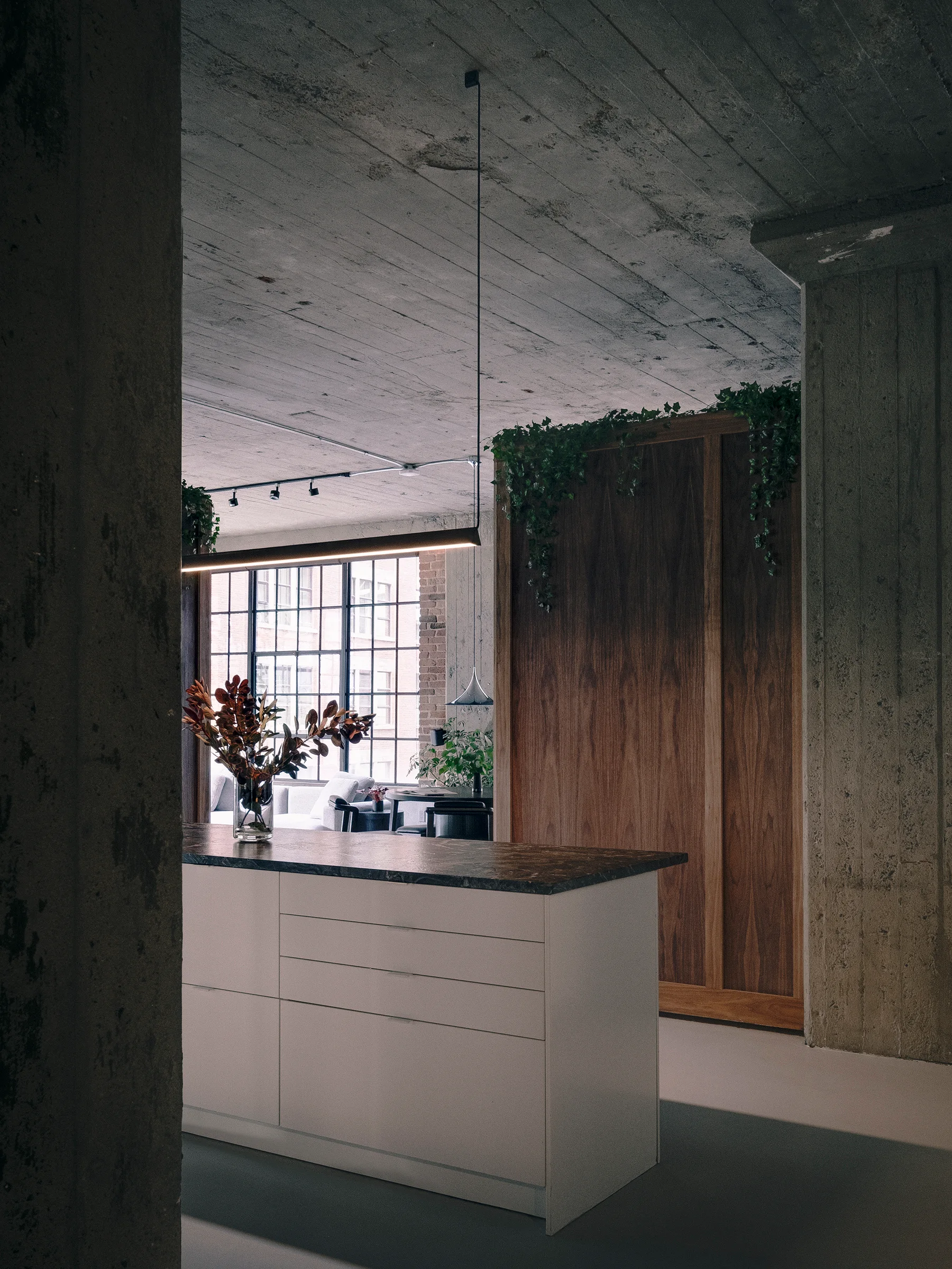 A modern loft kitchen features a white island with a dark countertop under an exposed concrete ceiling, beside a tall wood storage cabinet.