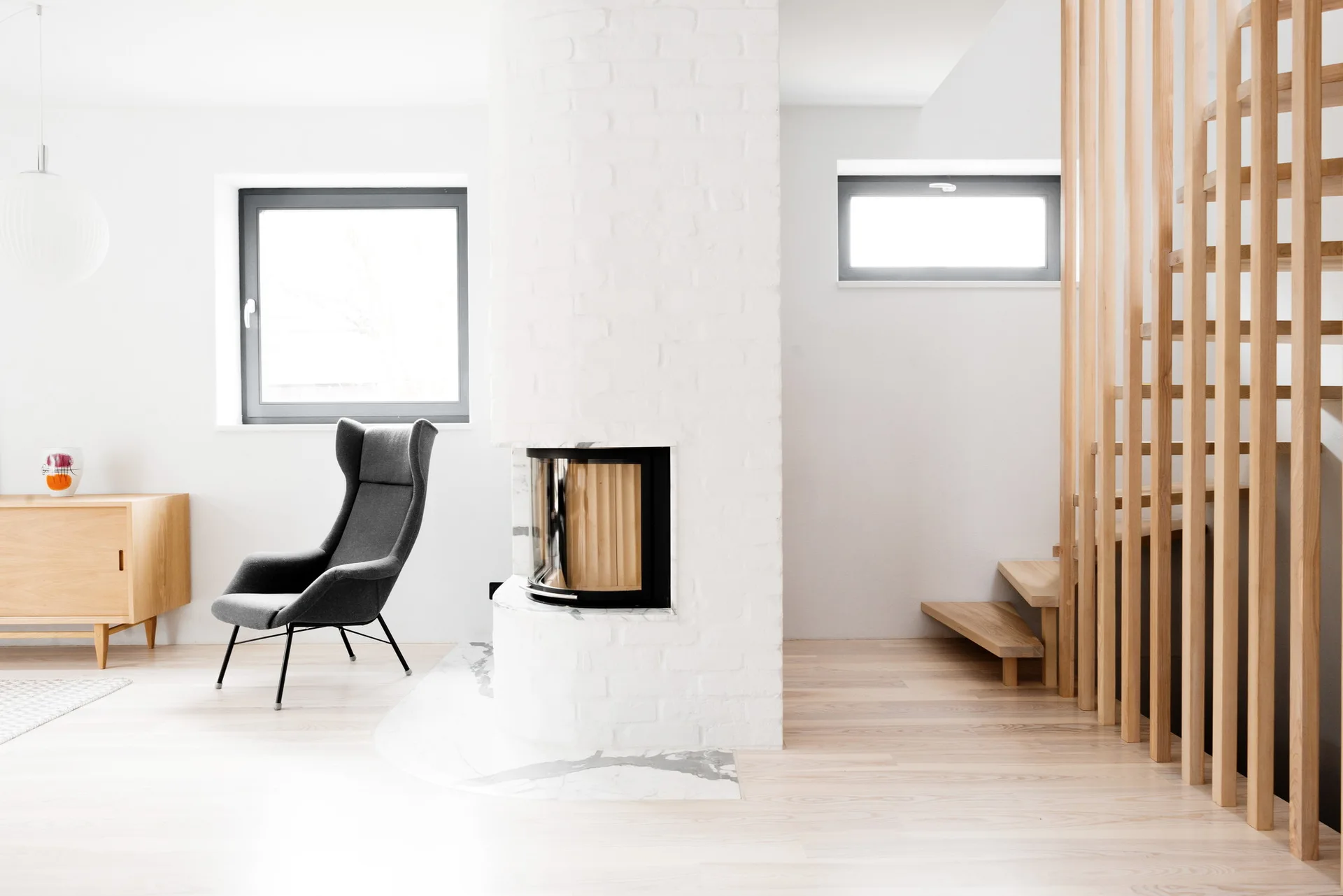 A minimalist room features a white painted brick fireplace partition, a dark gray armchair, and a light wood staircase with vertical slat railings.