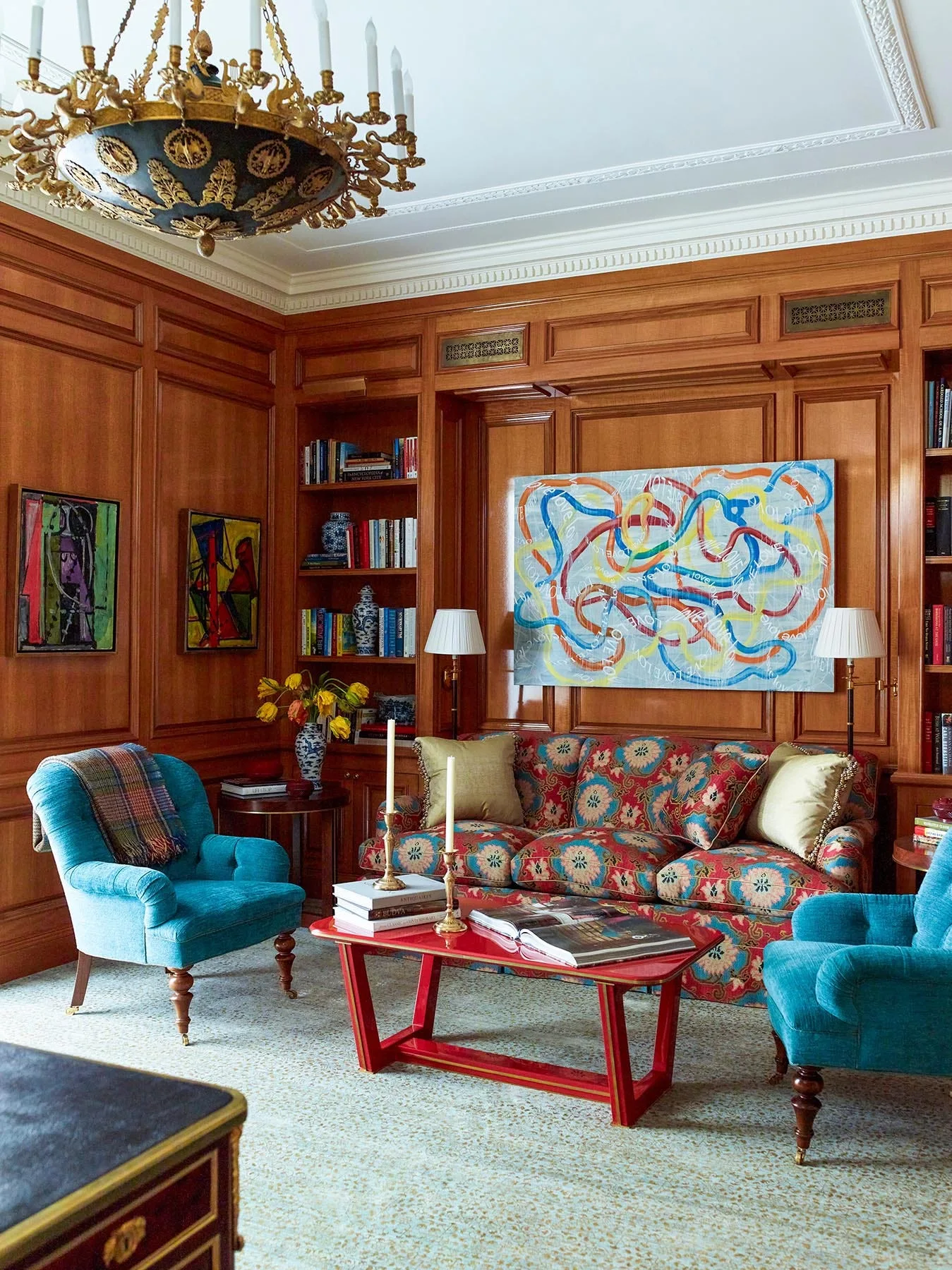 A traditional wood-paneled library features a floral-patterned sofa, teal armchairs, and a bright red coffee table under an ornate brass chandelier.