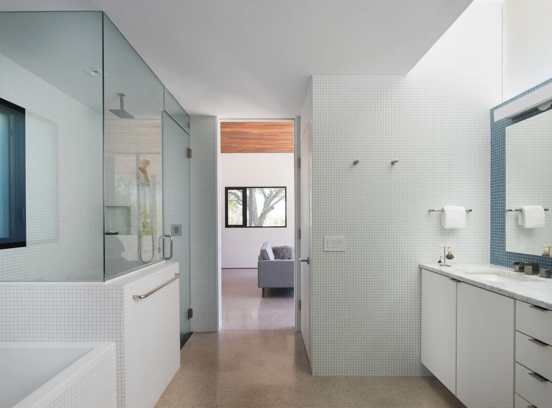 A contemporary bathroom featuring white mosaic tiles, a glass-enclosed shower, a white vanity with a marble counter, and a skylight.