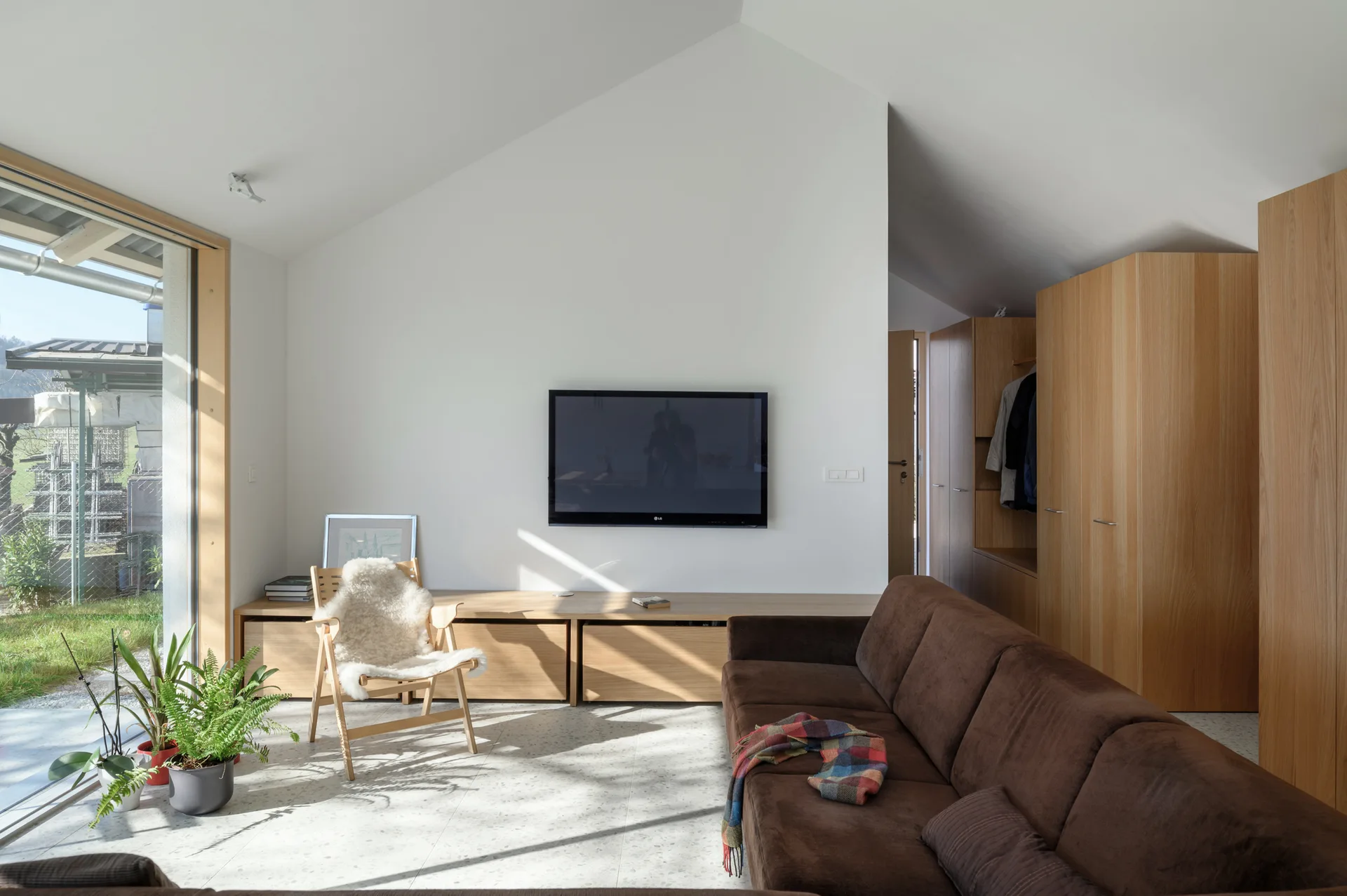 A minimalist living room featuring a vaulted ceiling, a brown sofa, a low wooden media console, and speckled terrazzo flooring.