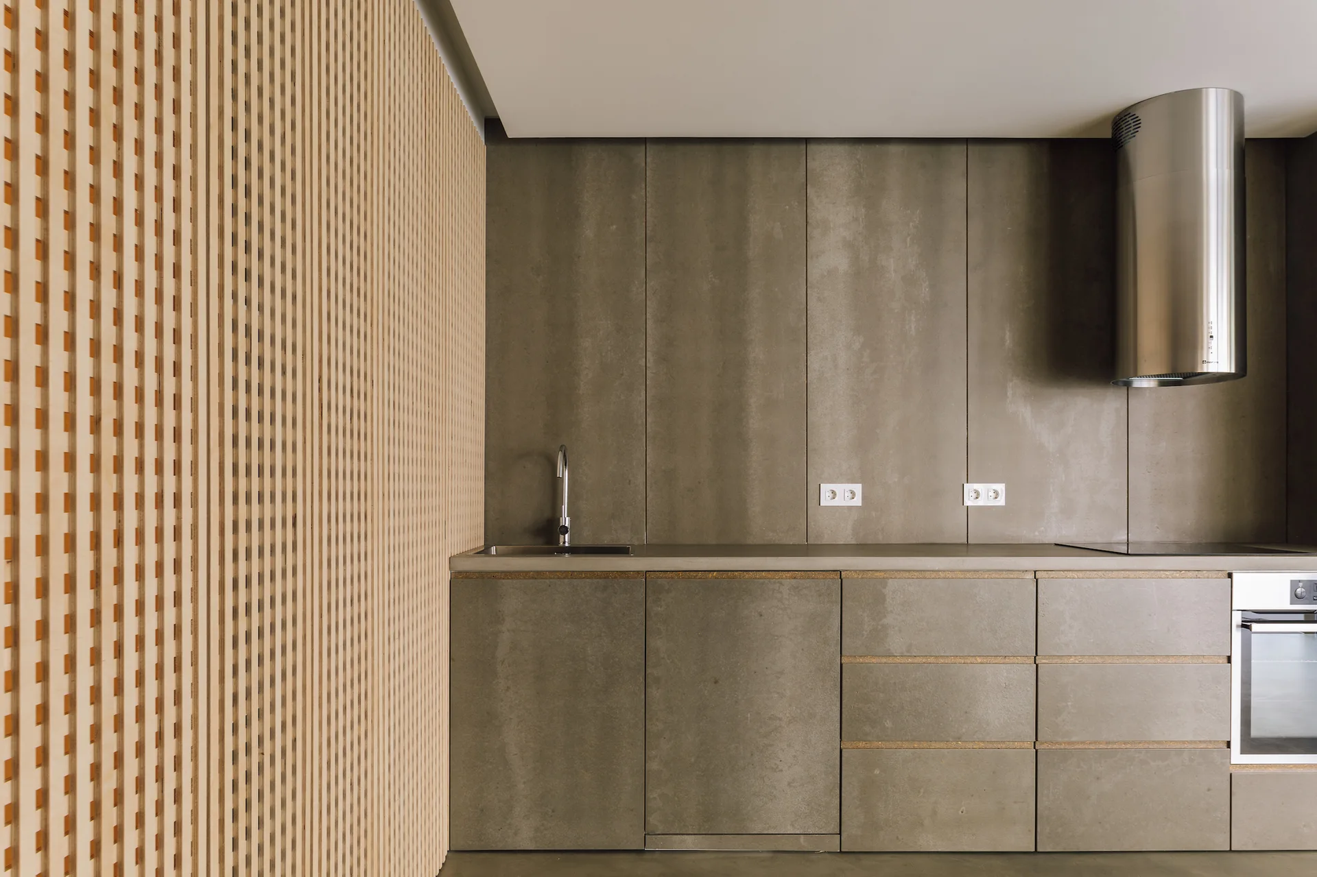A minimalist kitchen features concrete-look cabinets, a matching backsplash, a cylindrical range hood, and a light wood vertical slat wall on the left.