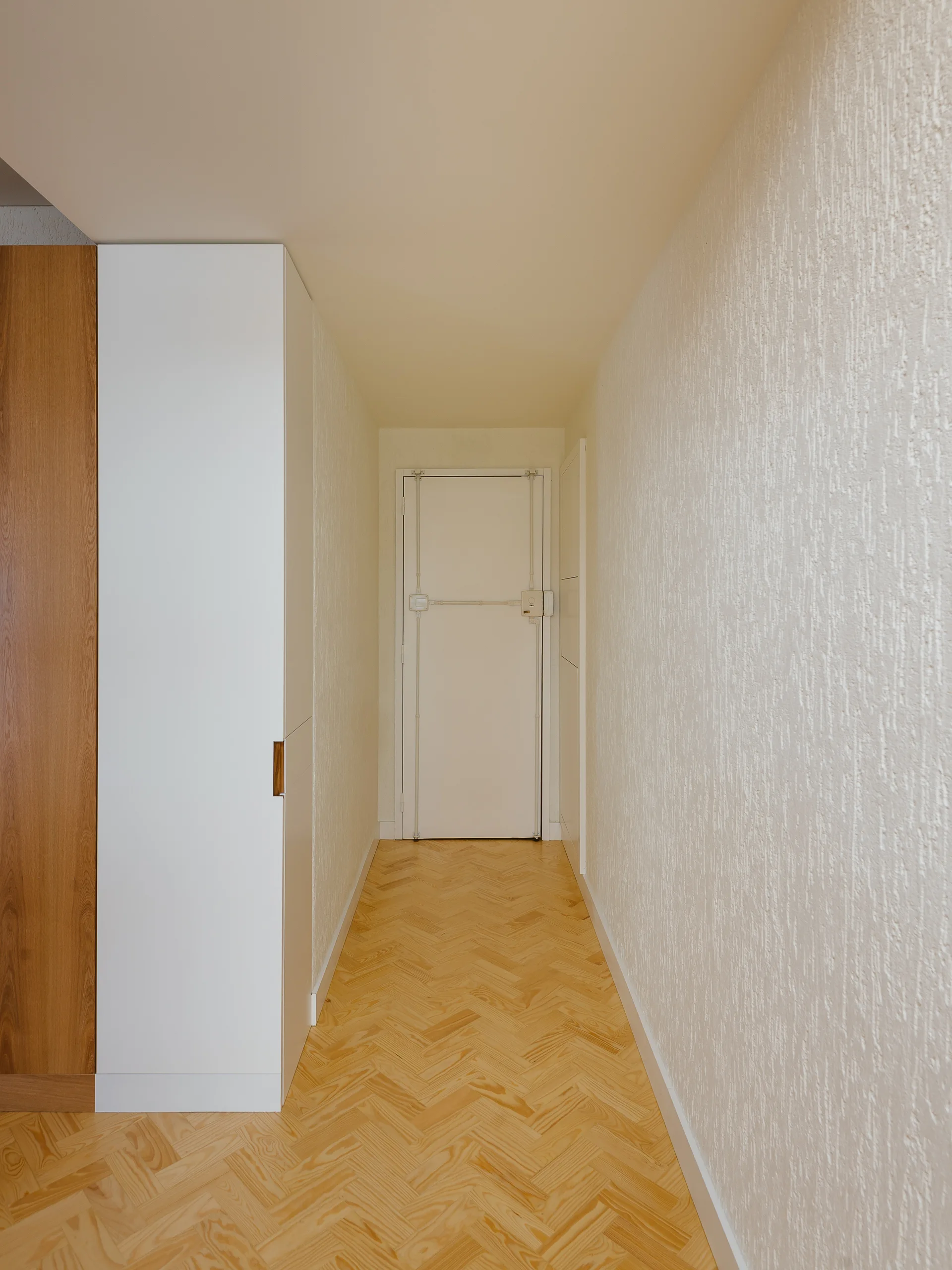 A narrow residential hallway features light wood herringbone flooring, heavily textured white walls, and a tall minimalist storage cabinet.