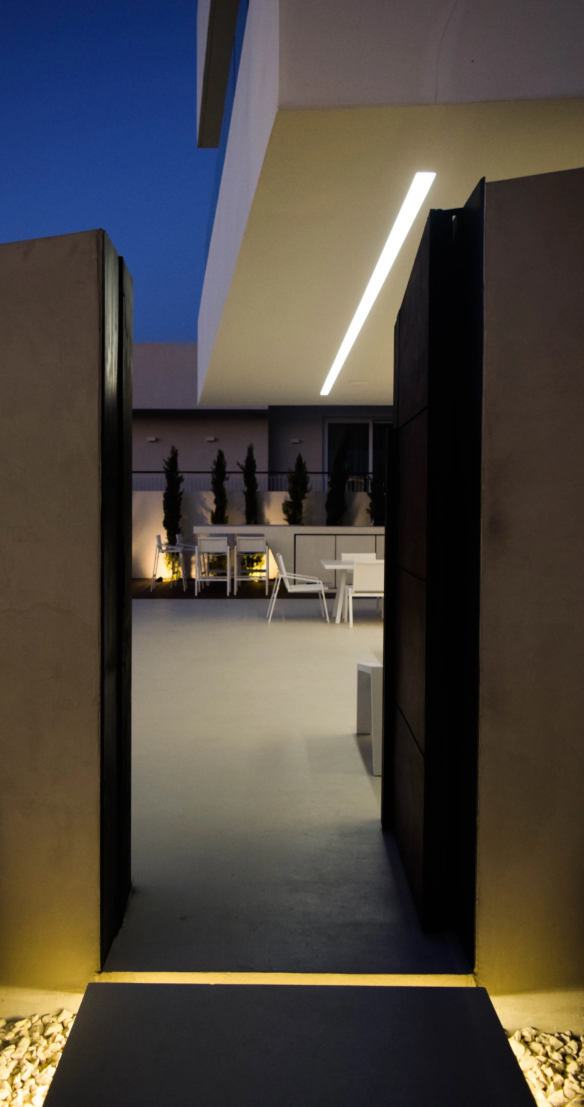 A concrete doorway looks out to a modern grey patio with white minimalist dining furniture and a glowing overhead linear light at dusk.