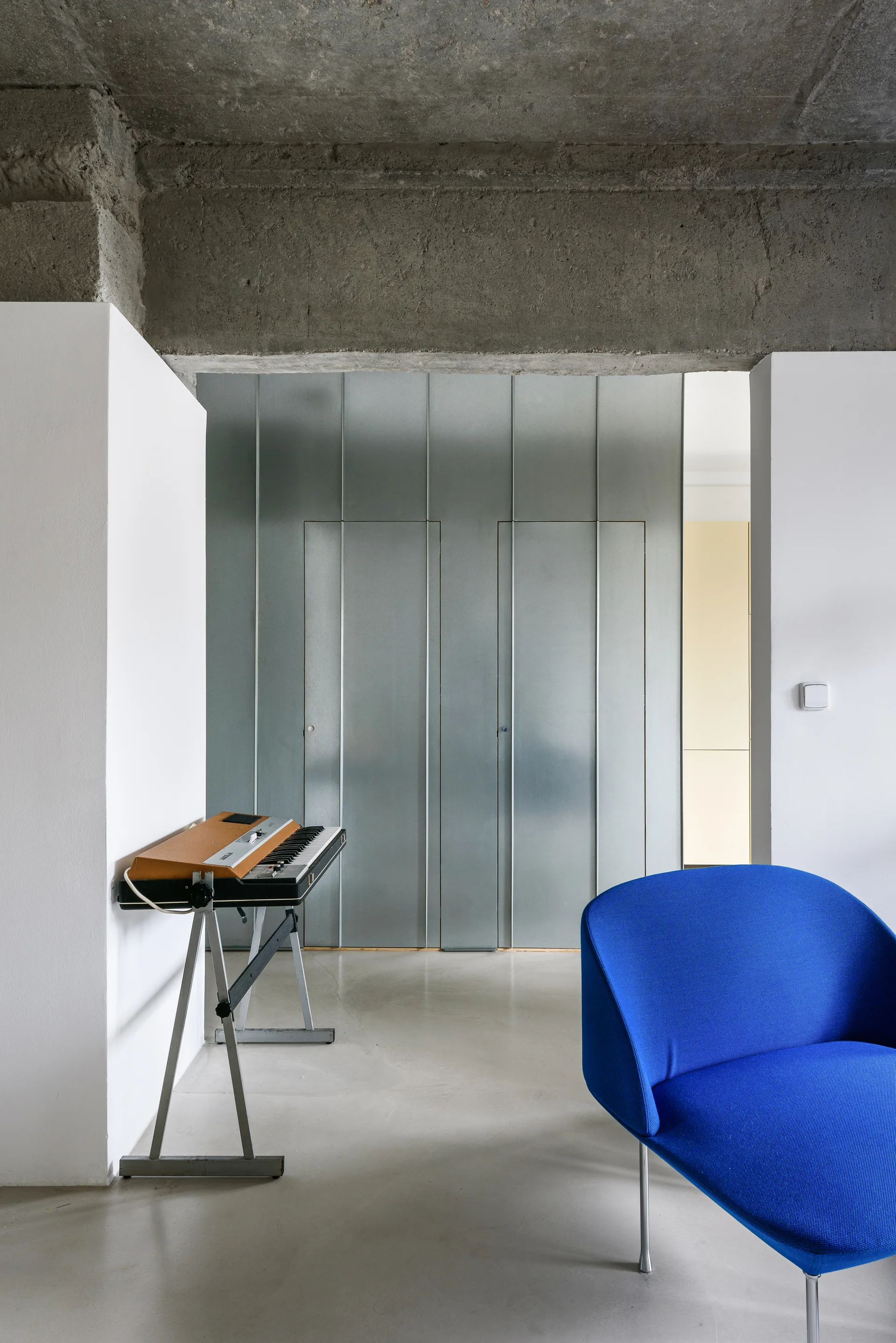 An electric blue lounge chair and vintage keyboard on a stand sit in a room with a raw concrete beam and metal wall panels.