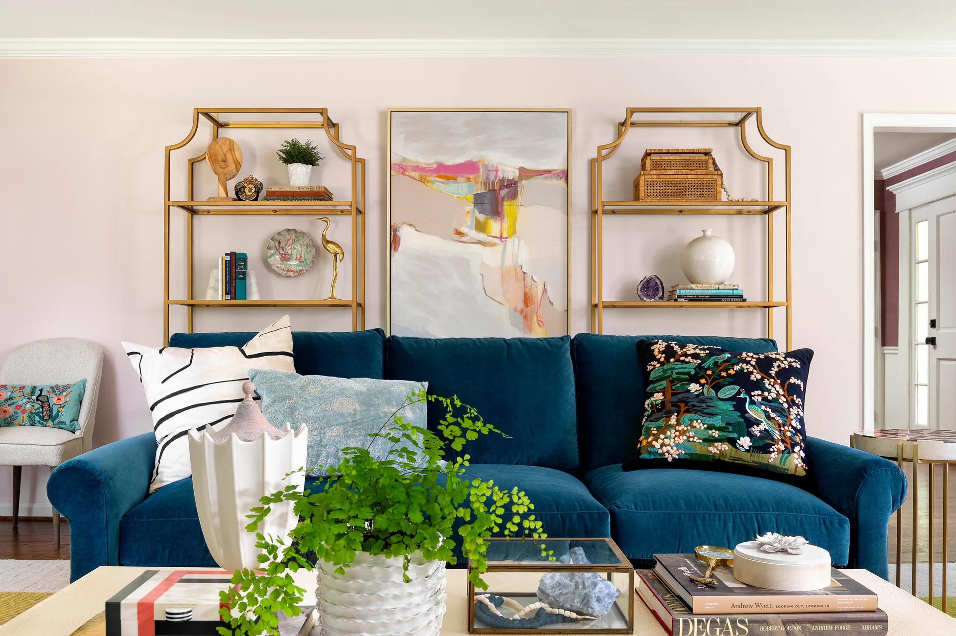 A blue velvet sofa sits between two brass arched shelving units against a pale pink wall beneath abstract art in a living room.