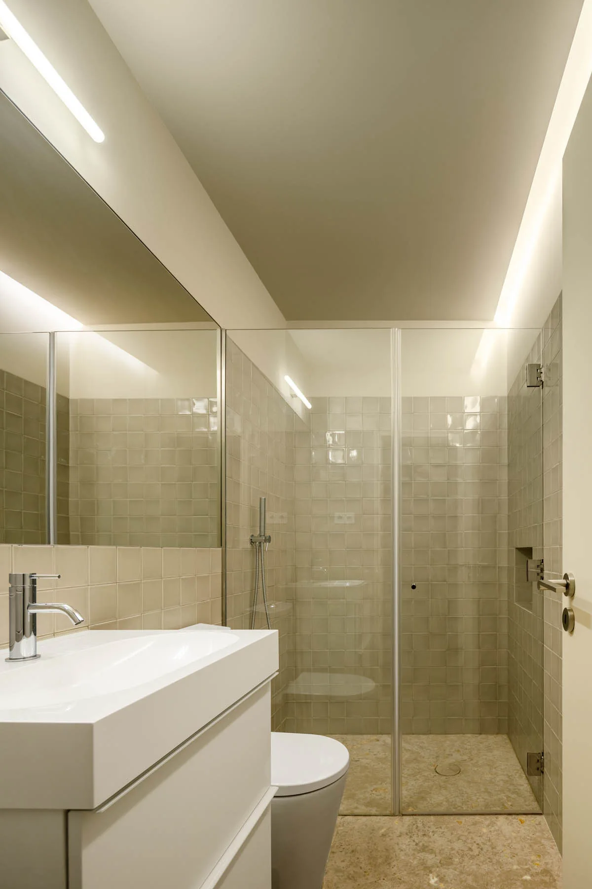 A modern bathroom features glossy beige square tiles on the wall, a white vanity sink, a frameless mirror, and a walk-in glass shower.