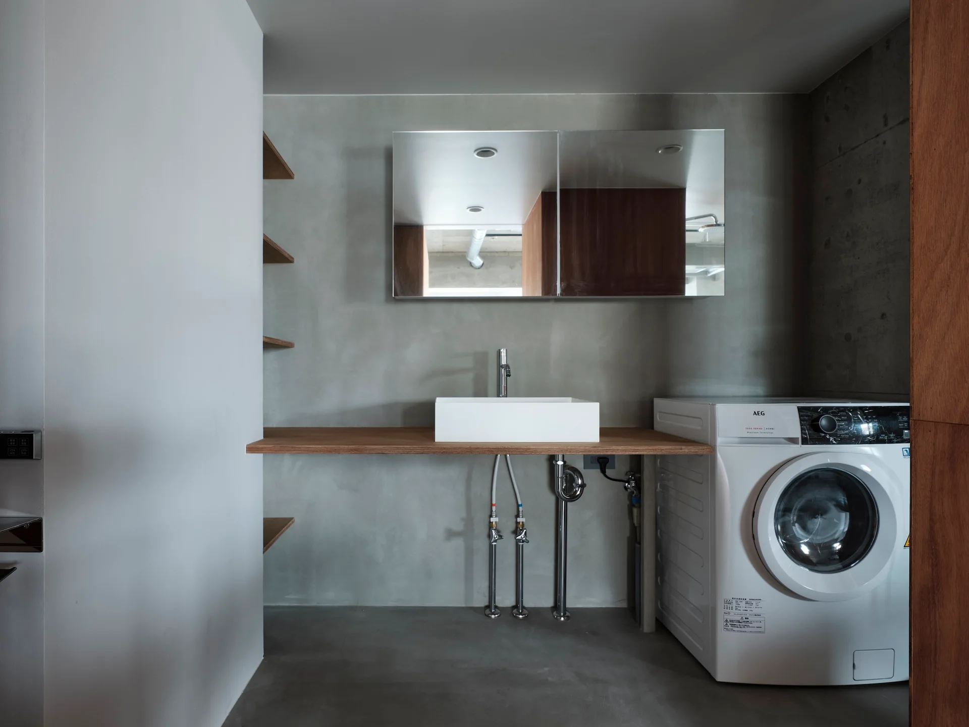 A minimalist laundry room featuring a white washing machine next to a rectangular vessel sink on a floating wooden counter against a concrete wall.