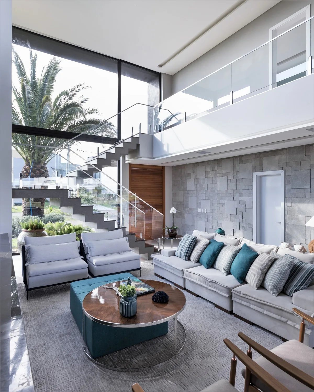 A modern double-height living room featuring a cream sectional sofa, a floating staircase with glass railings, and a gray stone accent wall.