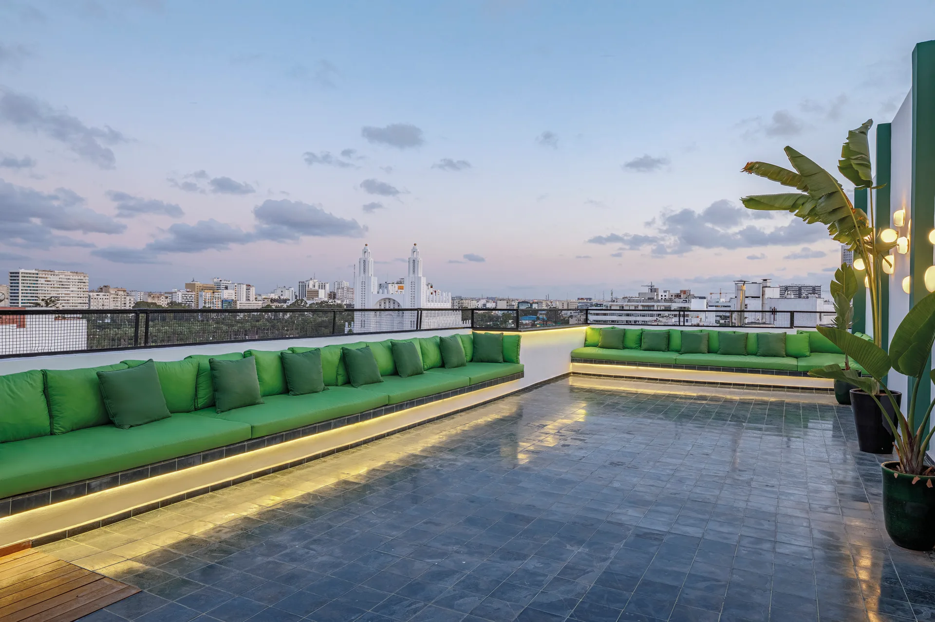 An outdoor rooftop terrace features long built-in benches with green cushions, dark grey tiled flooring, and under-bench LED lighting under a twilight sky.