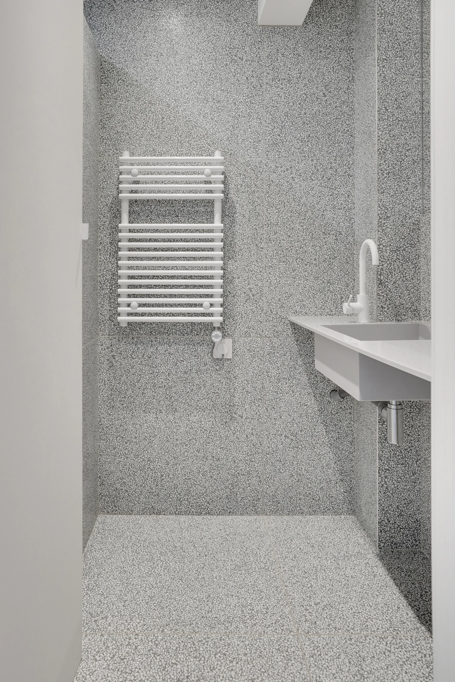 A minimalist bathroom features grey terrazzo-style wall and floor tiles, a white floating sink with a matching faucet, and a white ladder towel warmer.