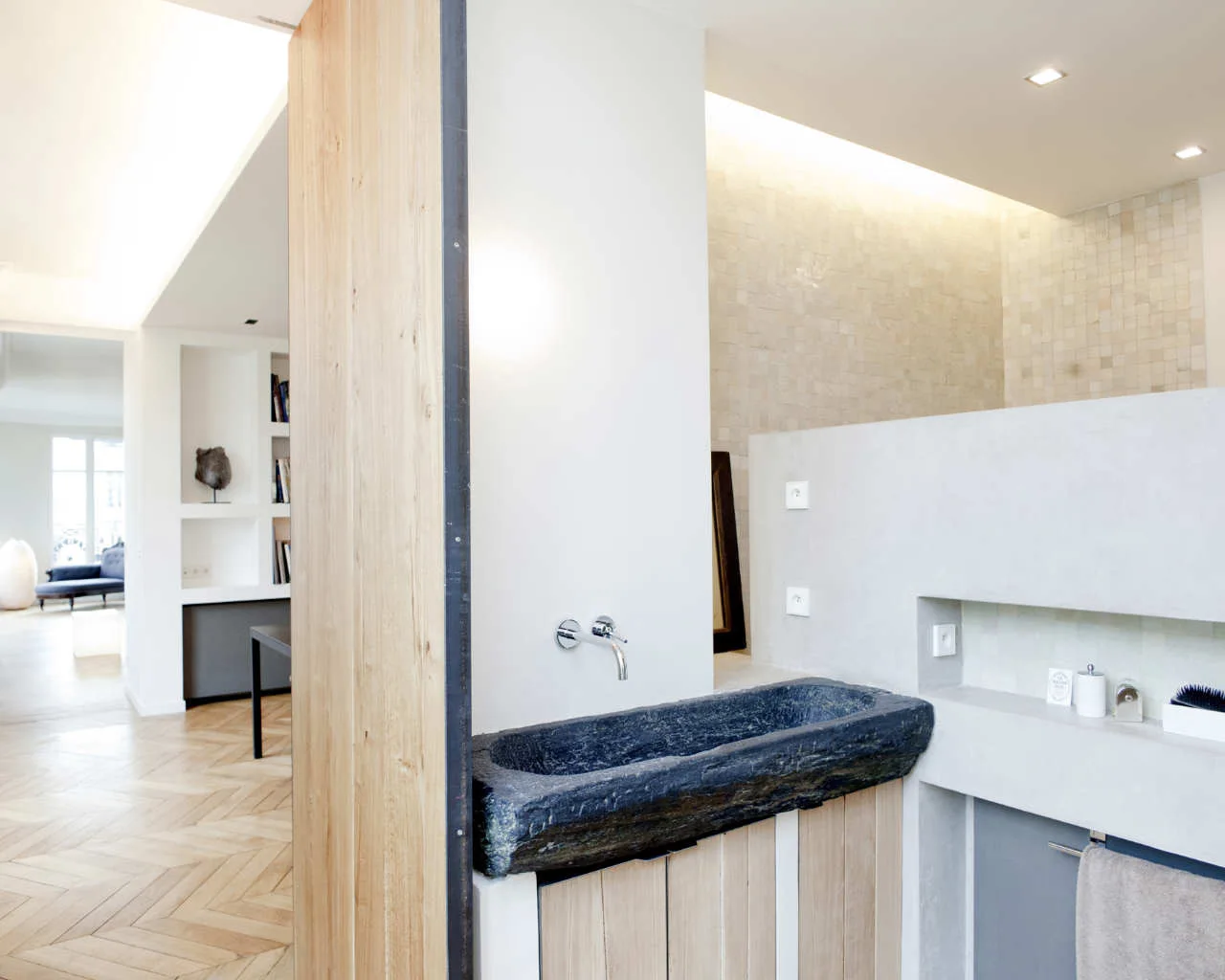 A modern bathroom featuring a rough-cut dark stone trough sink set on a light wood cabinet next to a smooth grey partition wall.