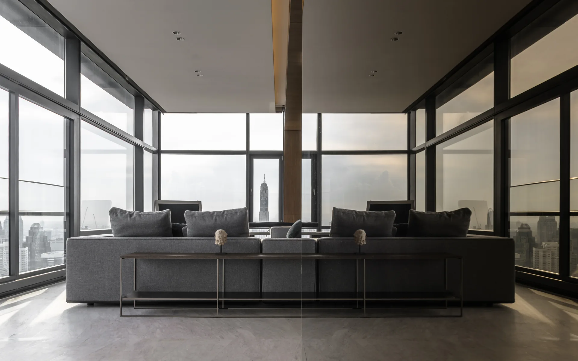 A minimalist living room features a long gray fabric sofa and a slim console table set against floor-to-ceiling windows showing a city skyline.