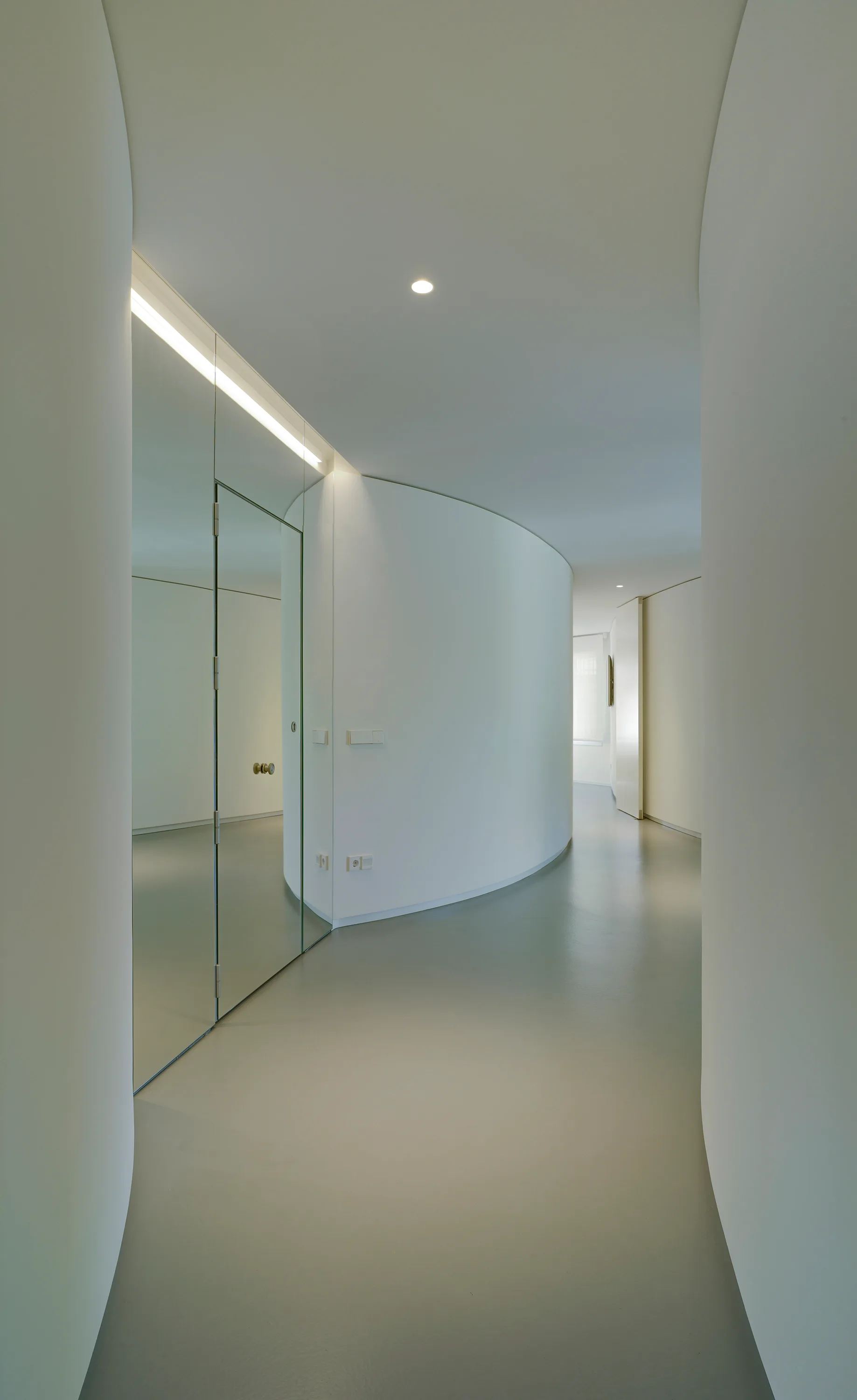 A minimalist hallway features a curved white plaster wall, seamless grey flooring, and a mirrored wall panel with an integrated flush door.