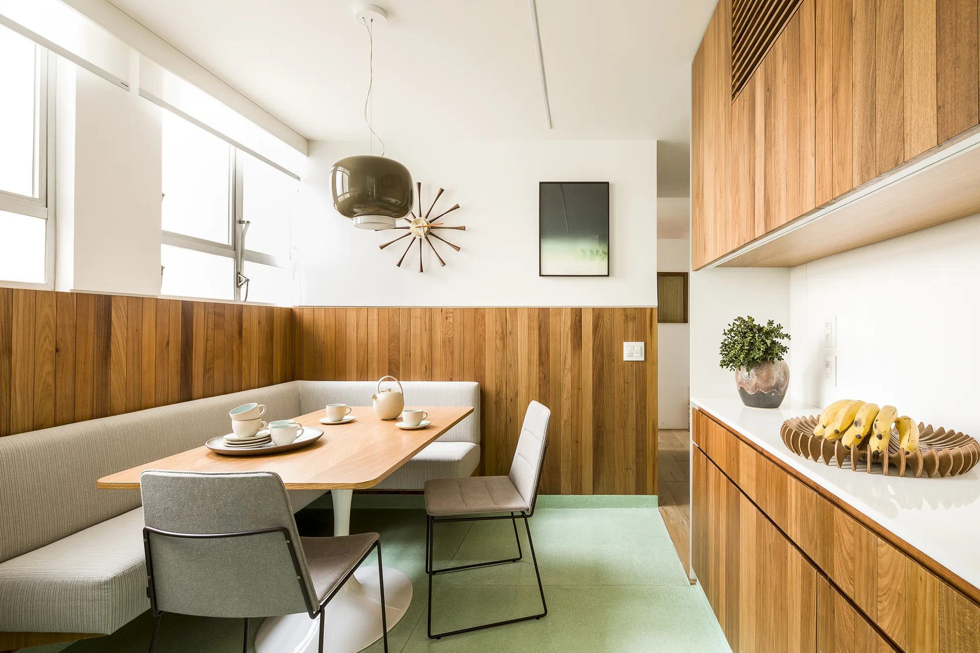 A mid-century modern dining nook features a built-in banquette, a pedestal table under a dome pendant light, and warm vertical wood wall paneling.