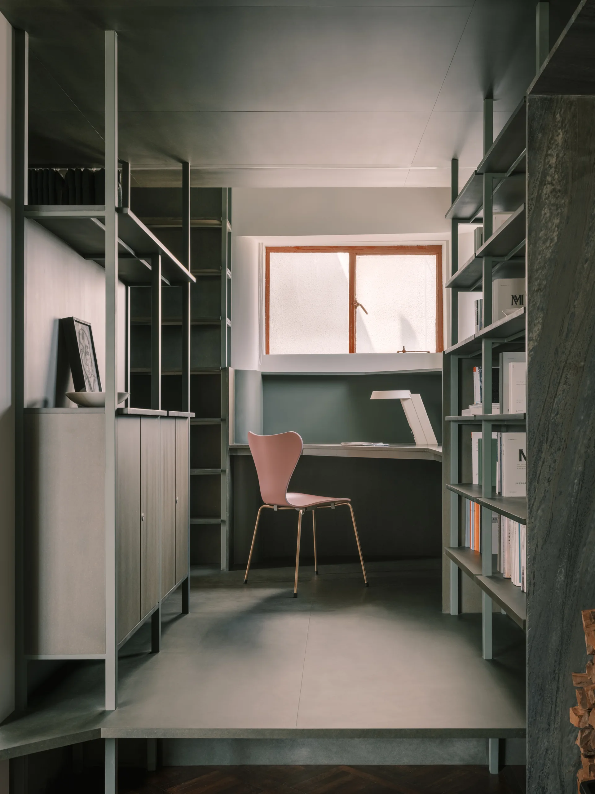 A minimalist home office with dark green walls and matching custom shelving features a built-in desk under a window and a muted pink chair.