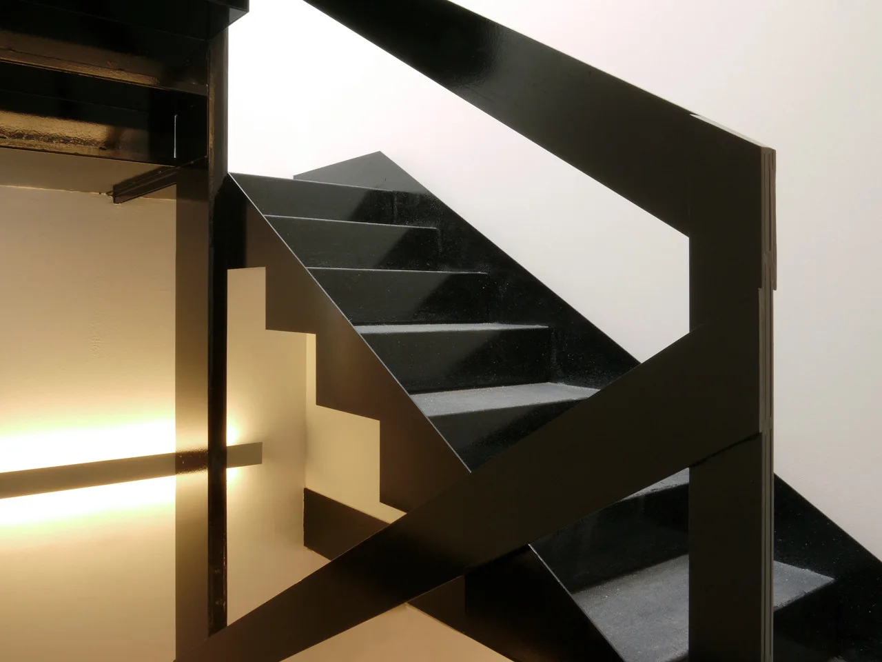 A modern black metal staircase with angular railings set against white walls and illuminated by a horizontal glowing strip light.