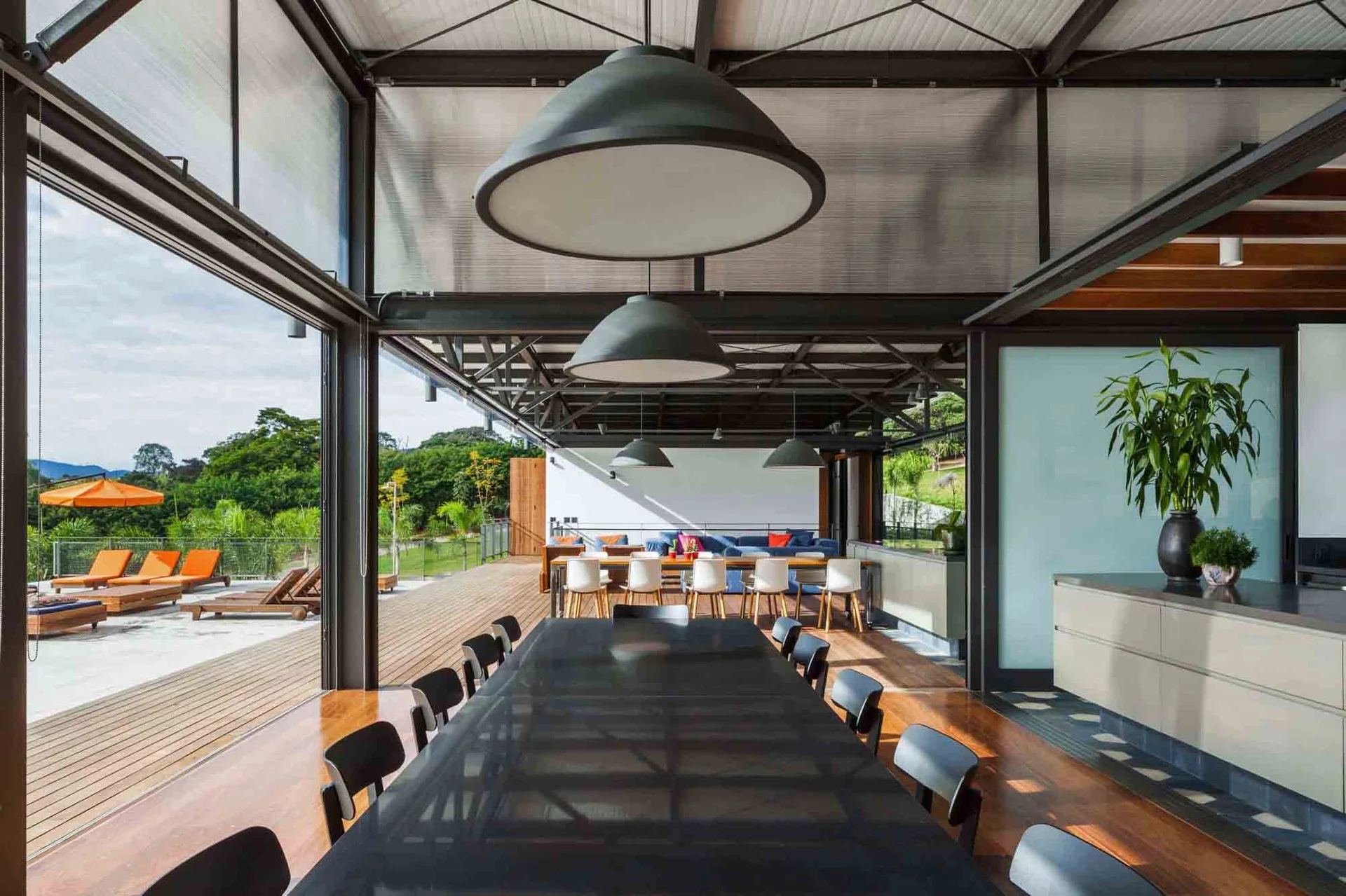 An open industrial dining room with metal ceiling trusses, large black pendant lights, and glass walls opening to an outdoor wooden deck.
