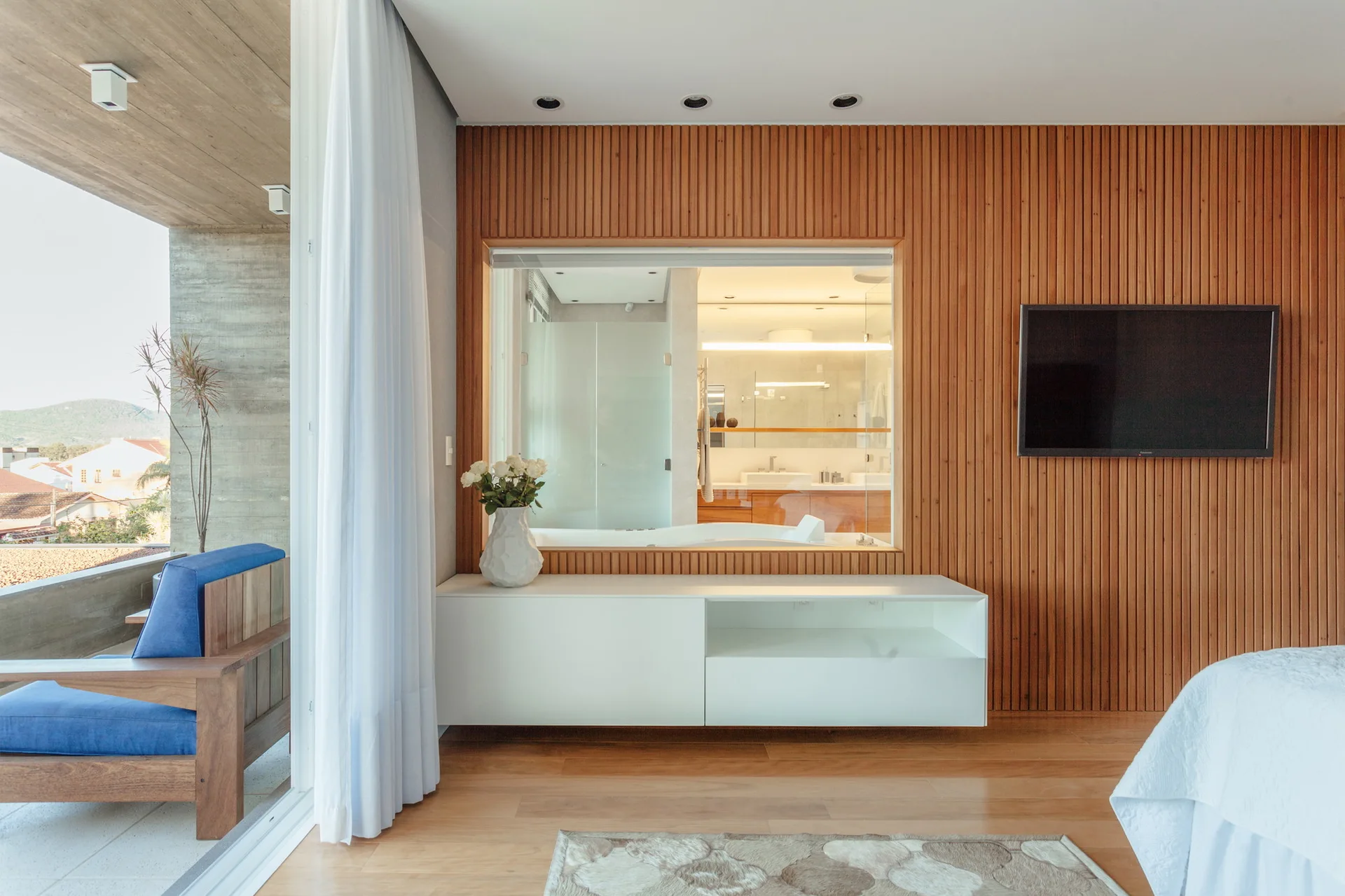 A modern bedroom with a vertical wood slat accent wall, a floating white media console, and an interior window looking into a bathroom.