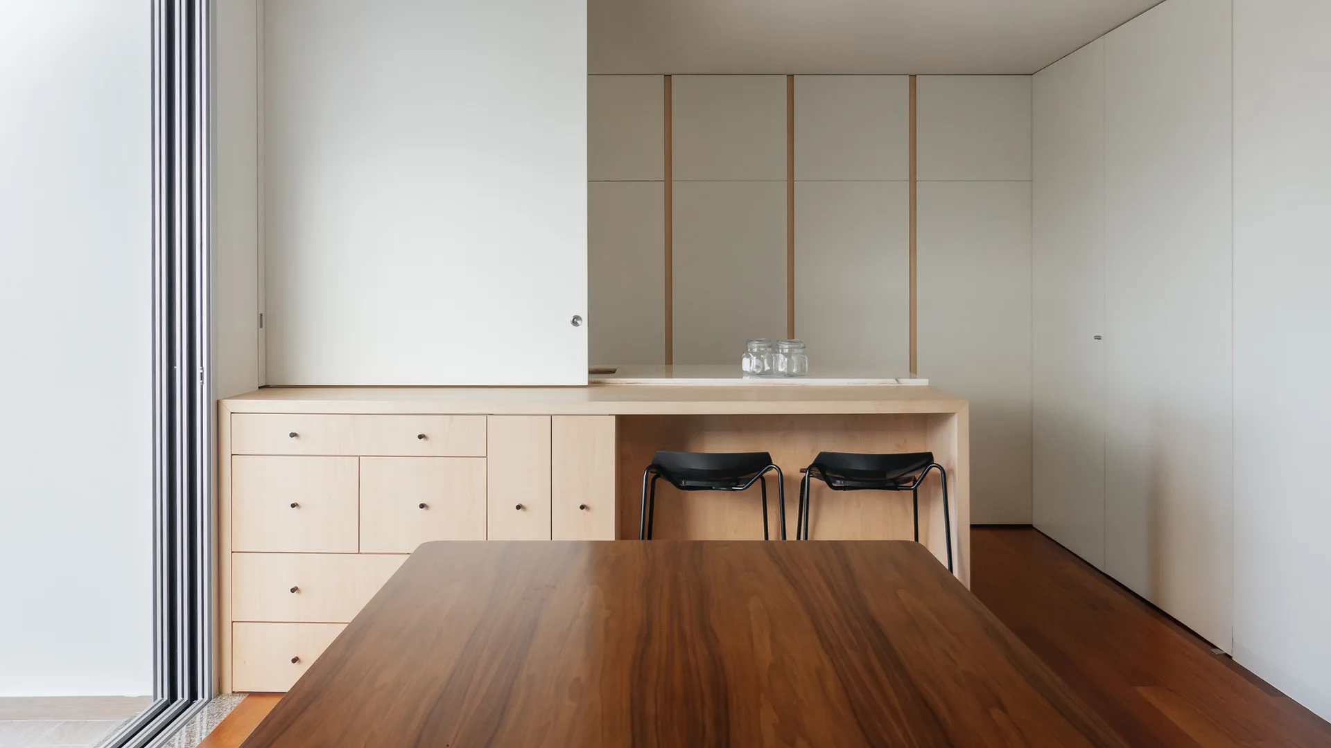 A minimalist kitchen features a dark wood dining table in the foreground, a light wood island with black barstools, and white wall cabinets.