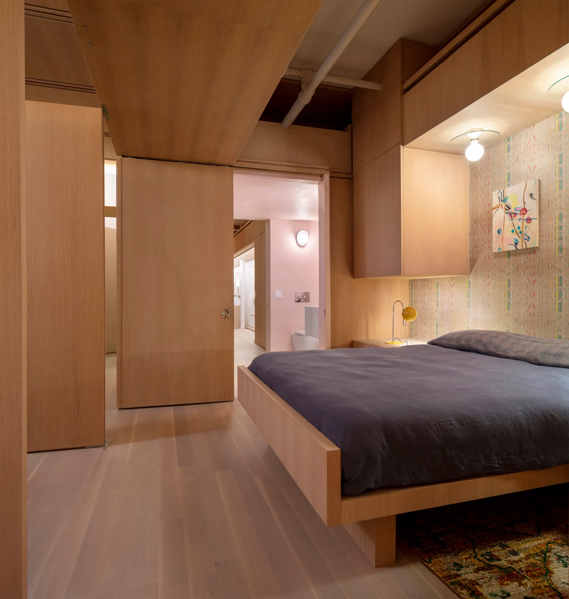 A modern bedroom features extensive light wood paneling, built-in cabinetry over a low-profile bed, and a sliding door opening to a pink hallway.